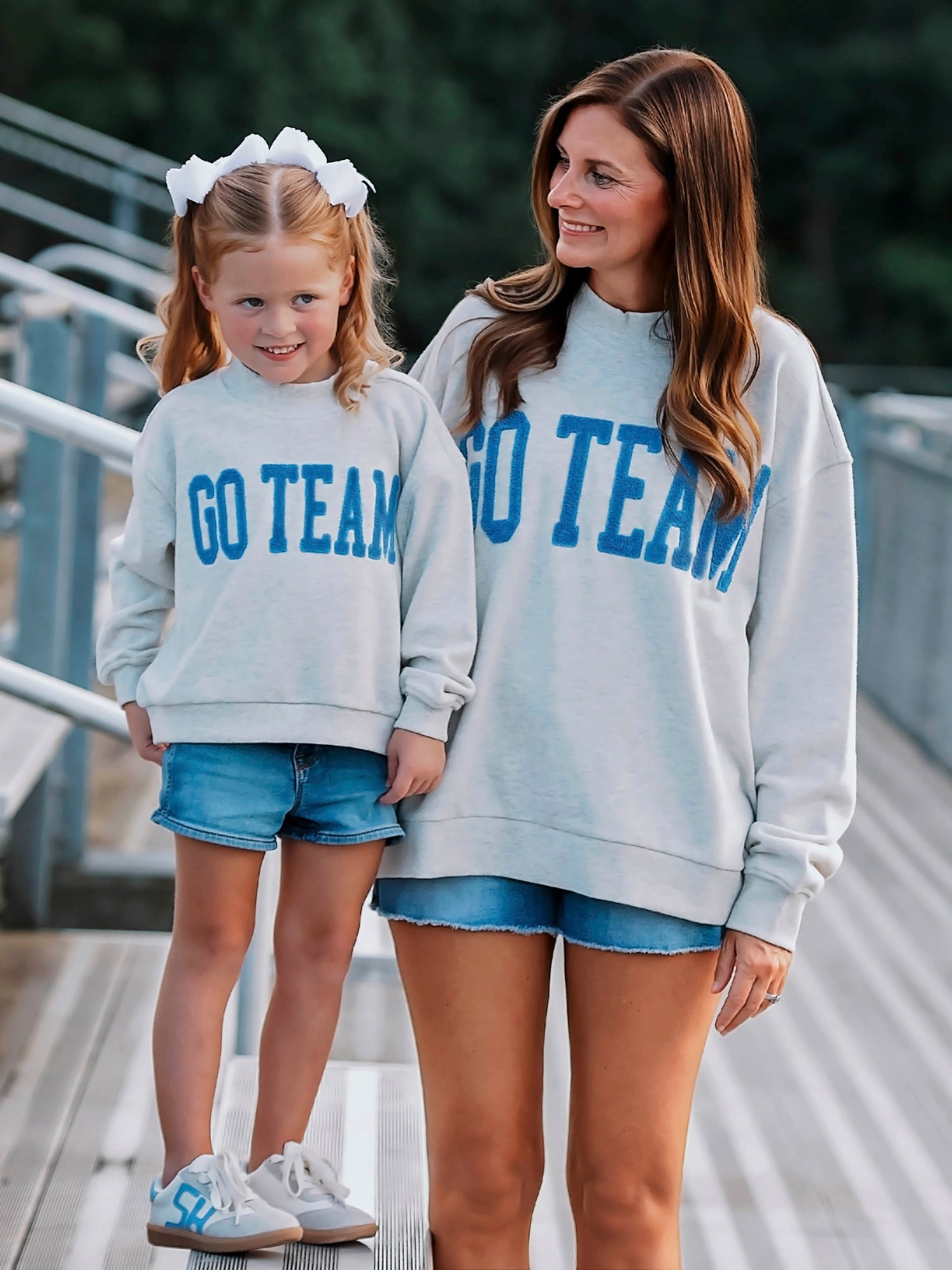 Women and child both wearing heathered gray sweatshirts that say GO TEAM in royal blue. Both appear to be standing on bleachers. 