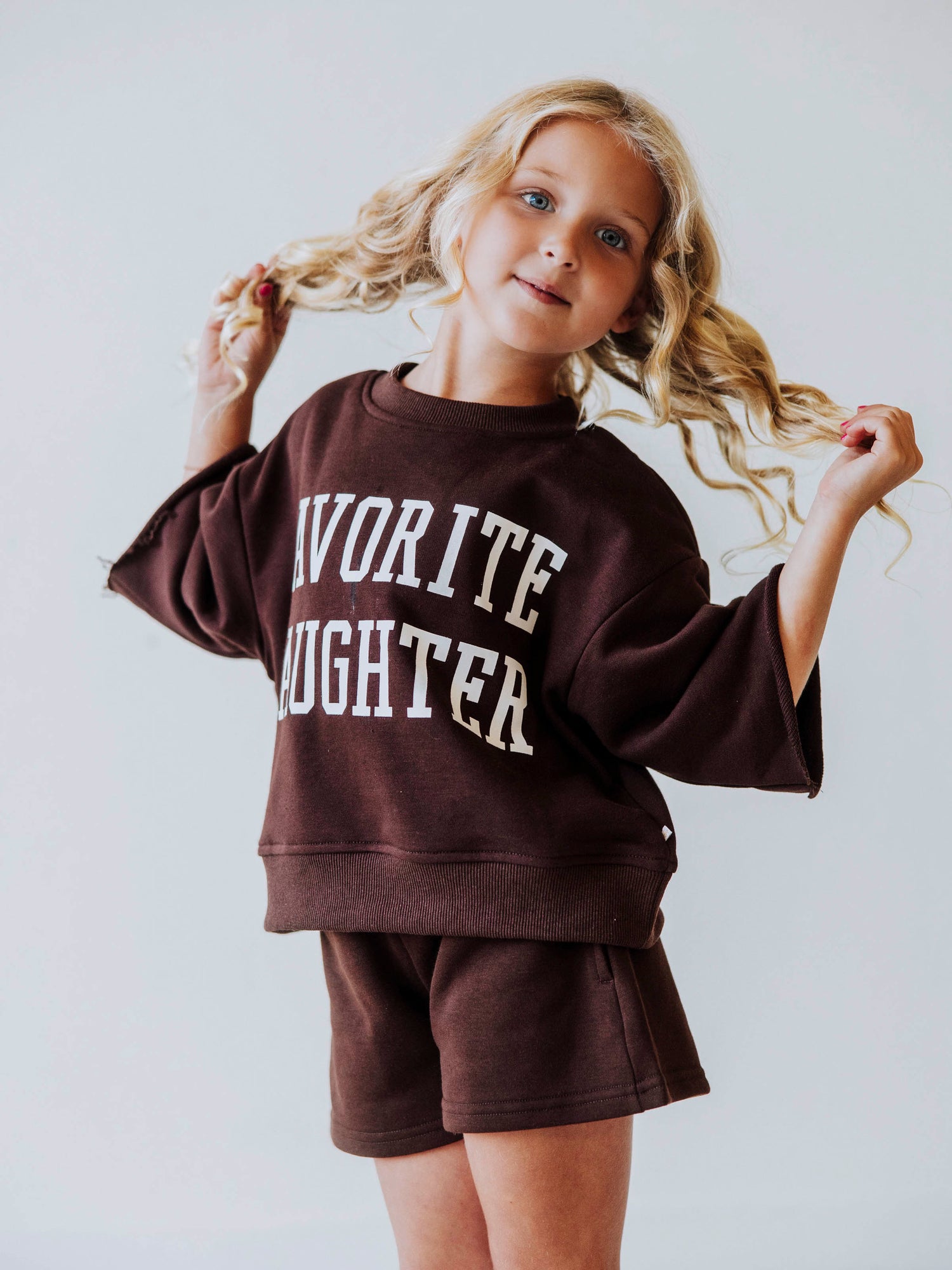Model wearing a two piece chocolate colored jogging set. Top says FAVORITE DAIGHTER. Bell hem sleeves with chocolate colored shorts. 
