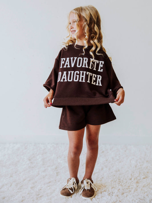 Model wearing a two piece chocolate colored jogging set. Top says FAVORITE DAUGHTER. Bell hem sleeves with chocolate colored shorts. 
