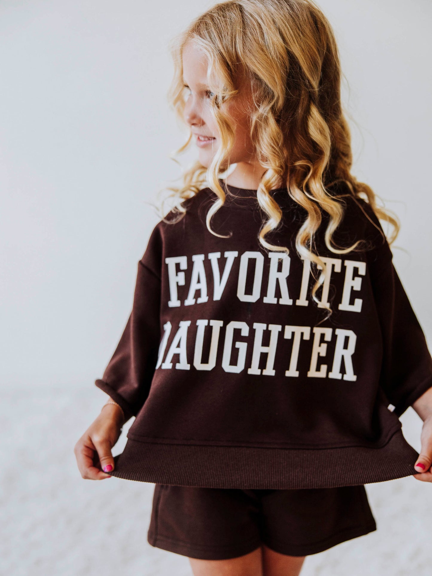 Model wearing a two piece chocolate colored jogging set. Top says FAVORITE DAIGHTER. Bell hem sleeves with chocolate colored shorts. 