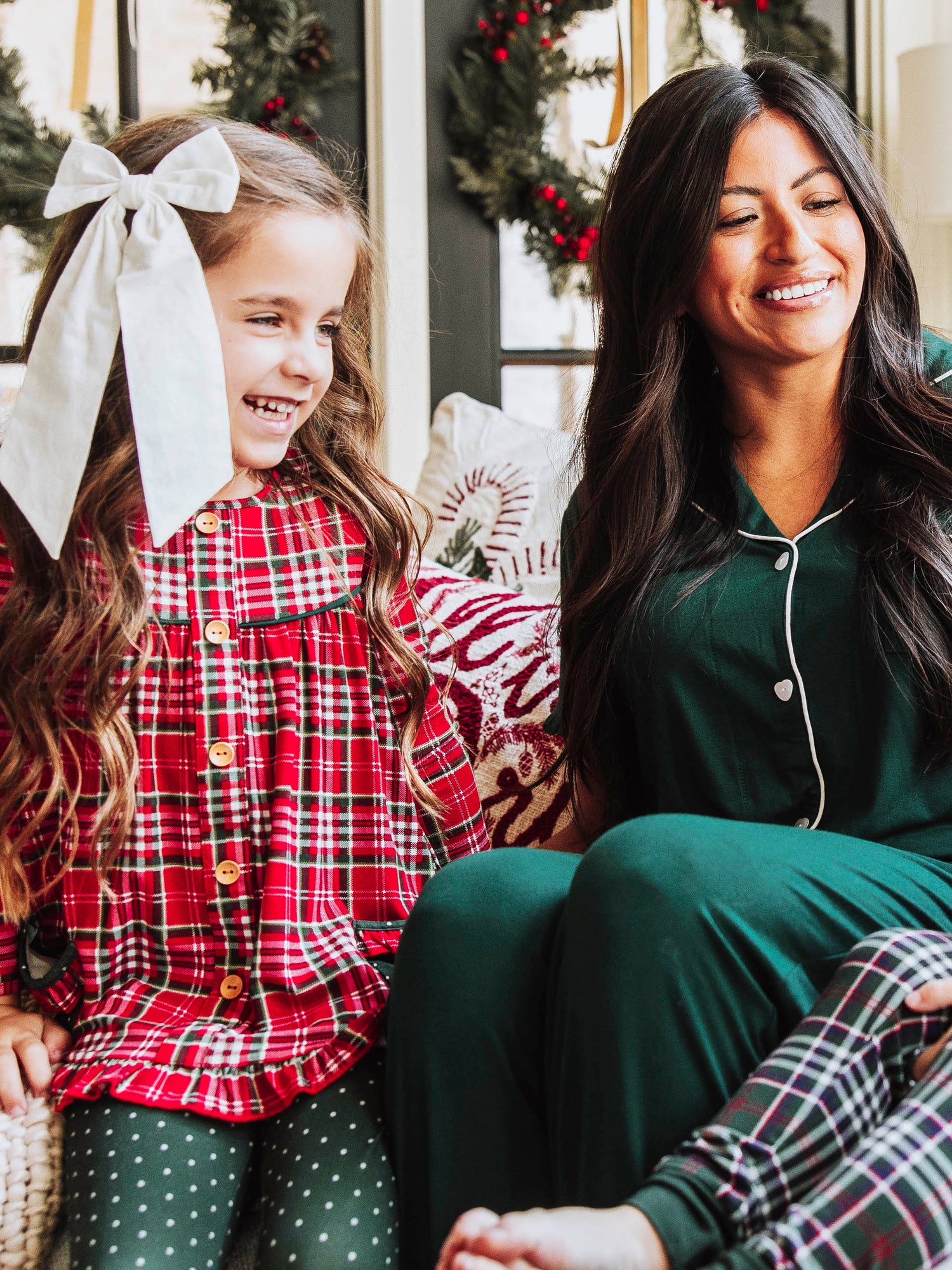 Girl and women both looking at another person maybe. Girl is wearing a traditional Christmas plaid top and has a bow in hair, women in wearing a deep green pajama set. 
