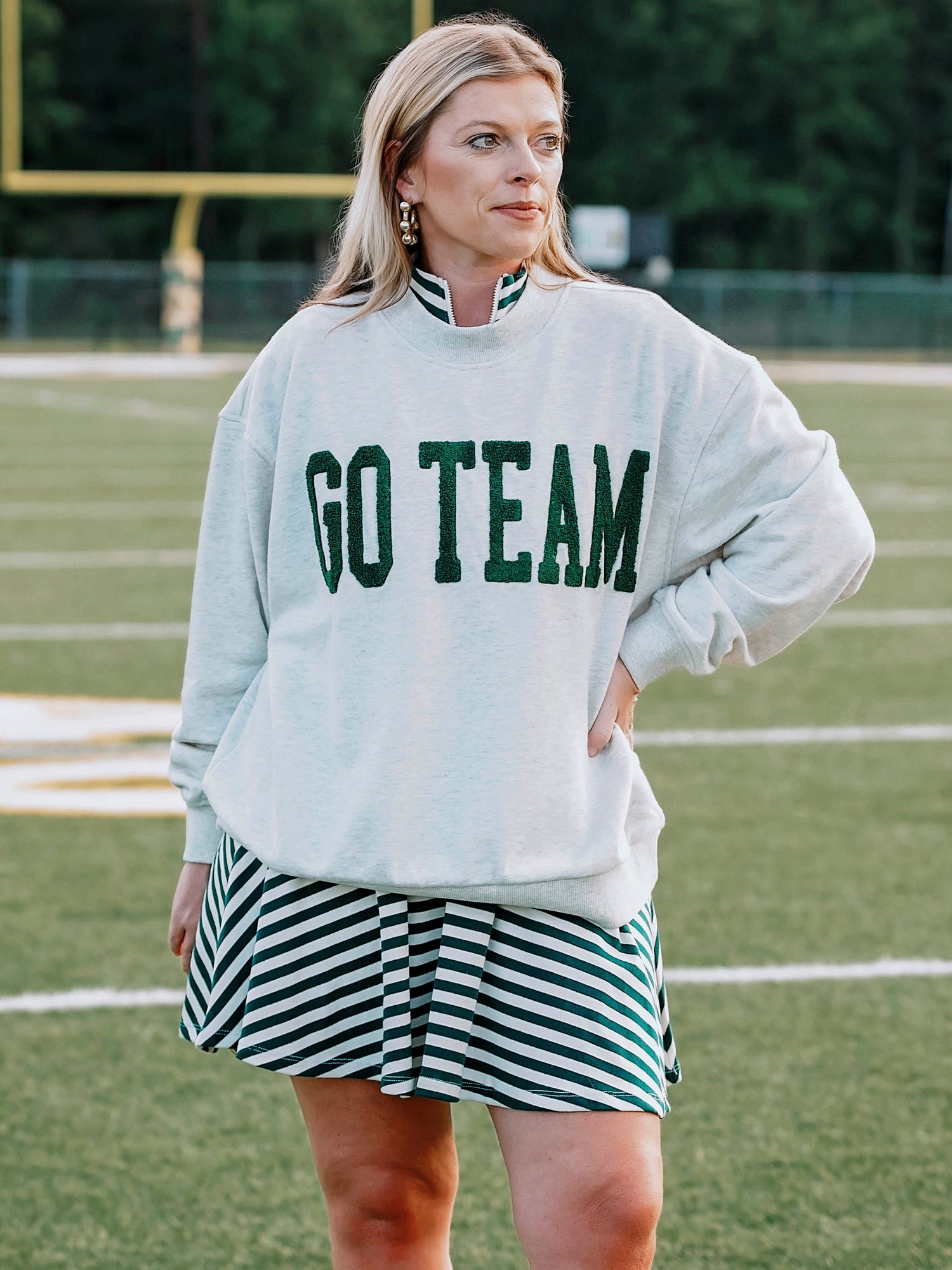 Women outside wearing a heathered gray sweatshirt that says GO TEAM in dark green. She is wearing the sweater over a green and white striped dress