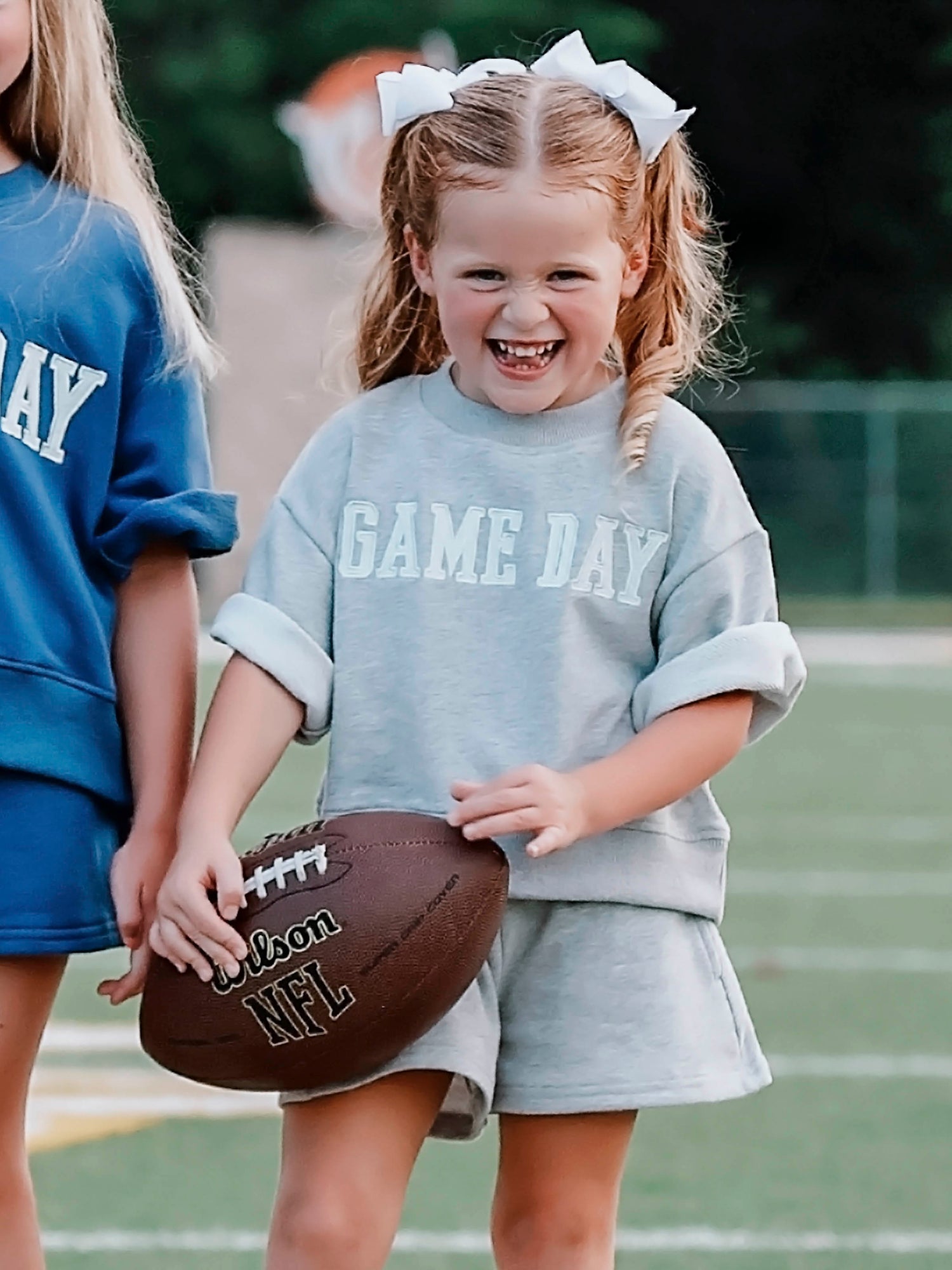 Girl outside wearing a two-piece gray sweat set that says GAME DAY on the front. Girl is holding a football.