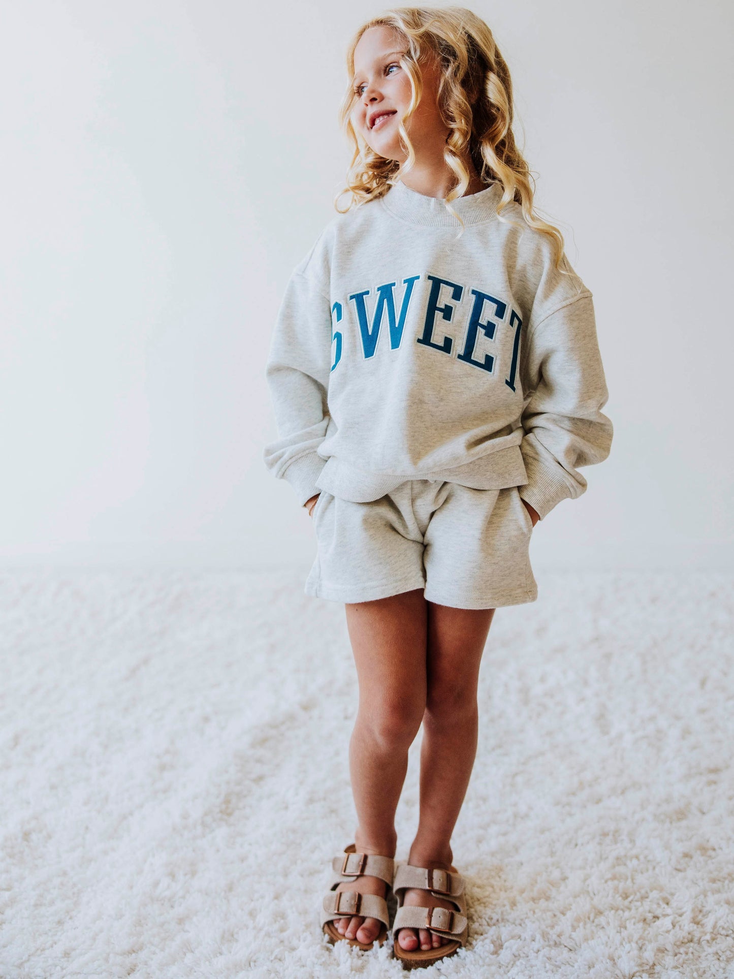 Girl model wearing a two piece gray in color set. The top says SWEET on the front in blue letters. Model is also wearing suede strapped sandals. Bottoms of set are same gray as top. 
