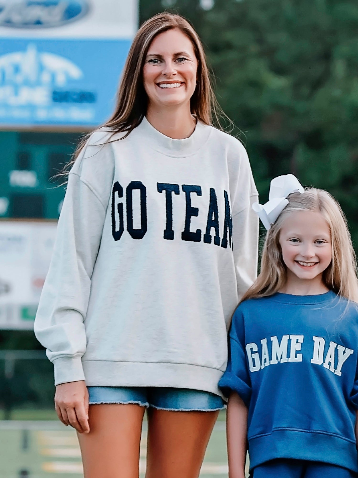 Women outside wearing a heathered gray sweater that says GO TEAM in a deep navy color. Also beside her is a little girl wearing a GAME DAY navy set