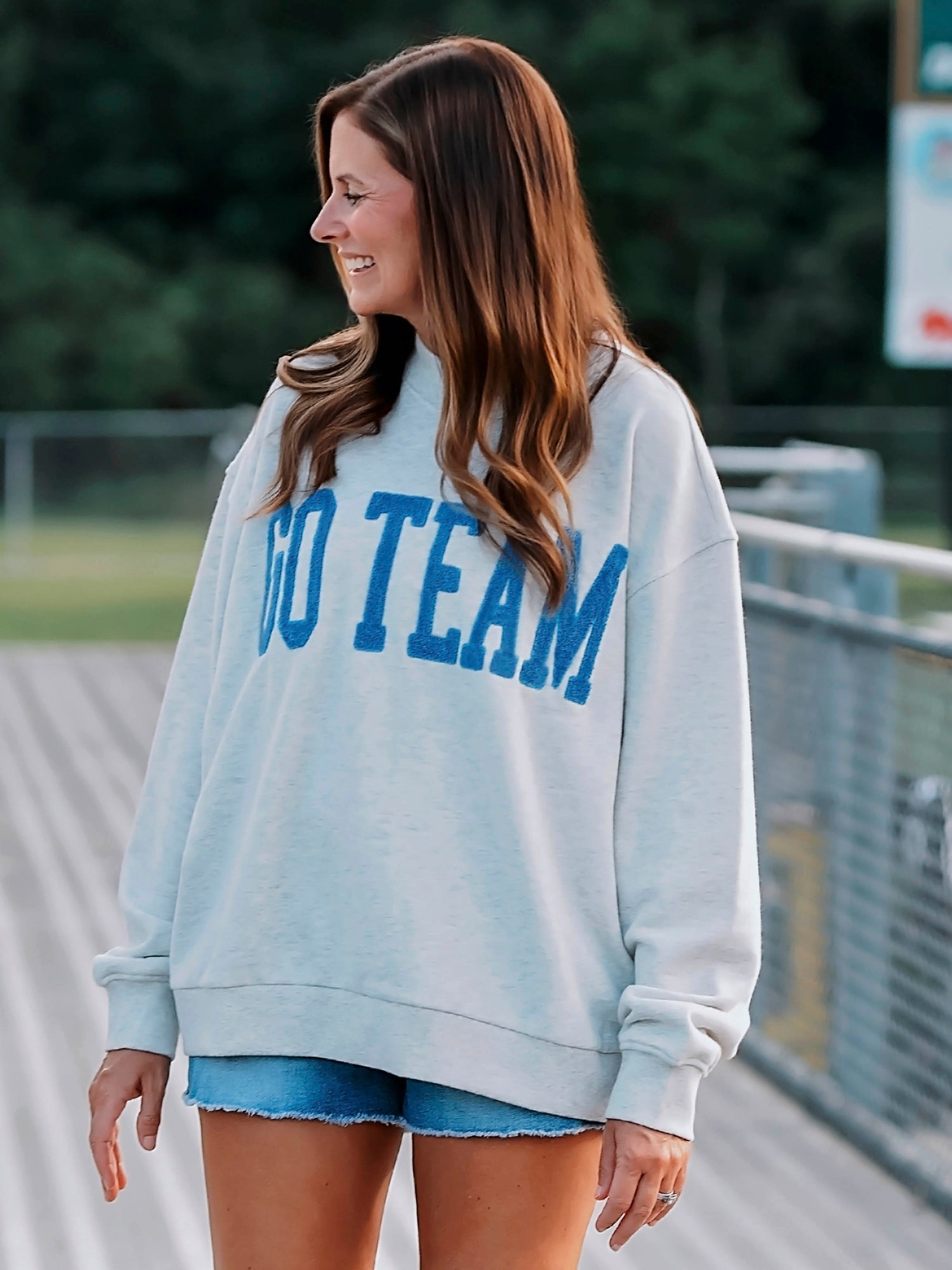 Women wearing a heathered gray sweatshirt that says GO TEAM on the front in royal blue. Women appears to be standing on bleachers. 