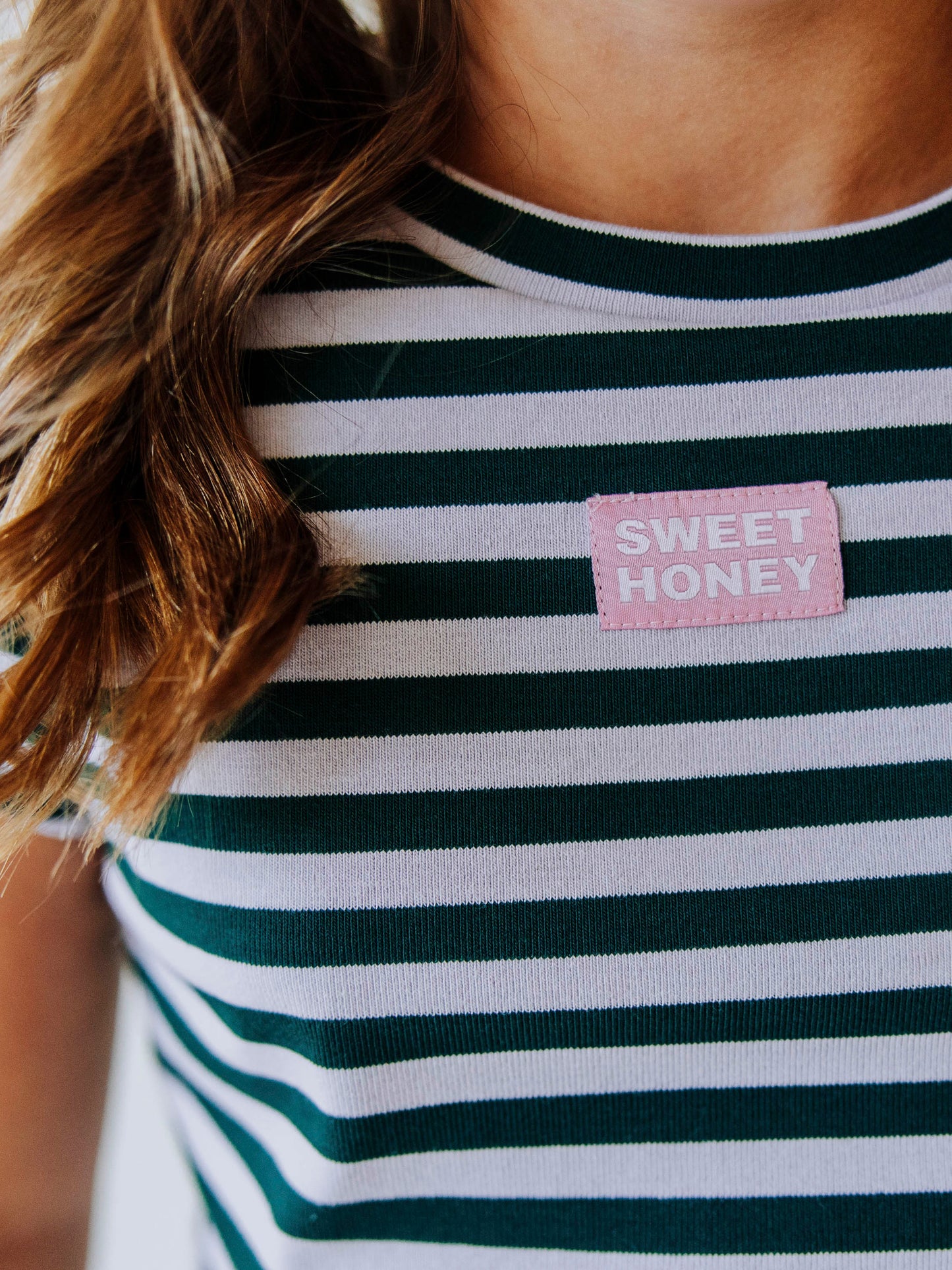 Up close shot of pink and white SweetHoney tag that is stitched on center of green and gray colored shirt
