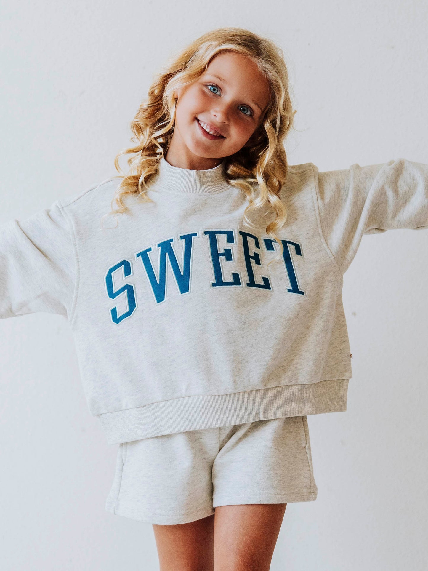 Girl model wearing a two piece gray in color set. The top says SWEET on the front in blue letters. Model is also wearing suede strapped sandals. Bottoms of set are same gray as top. 