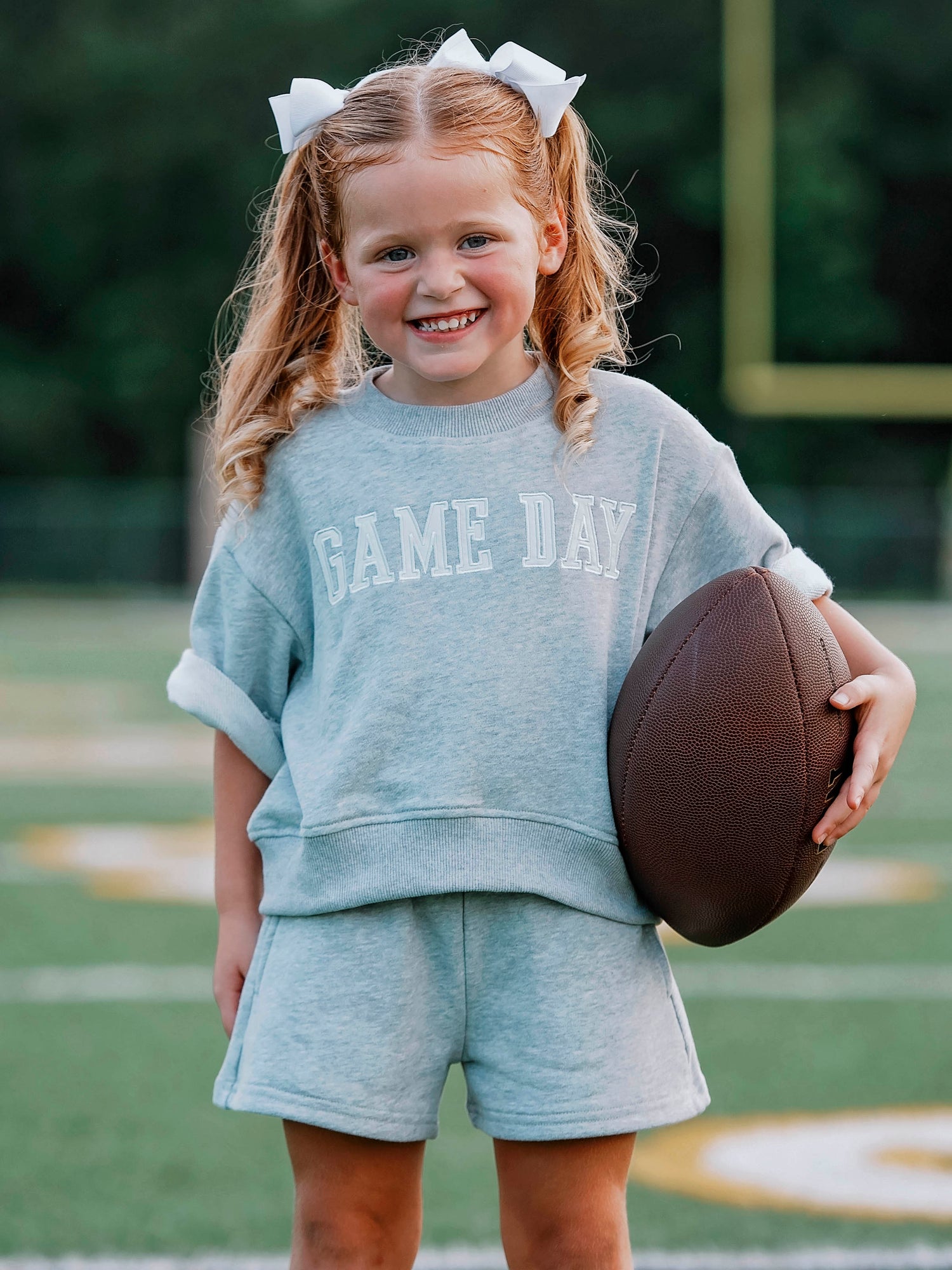 Girl outside wearing a two-piece gray sweat set that says GAME DAY on the front. Girl is holding a football.