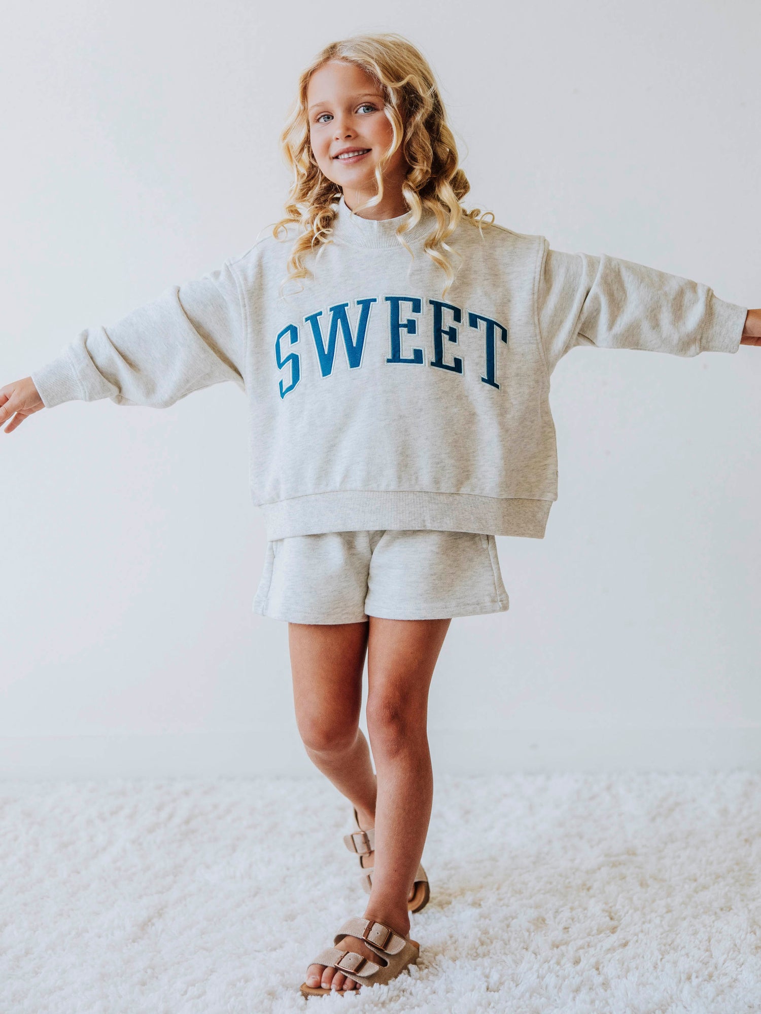 Girl model wearing a two piece gray in color set. The top says SWEET on the front in blue letters. Model is also wearing suede strapped sandals. Bottoms of set are same gray as top. 