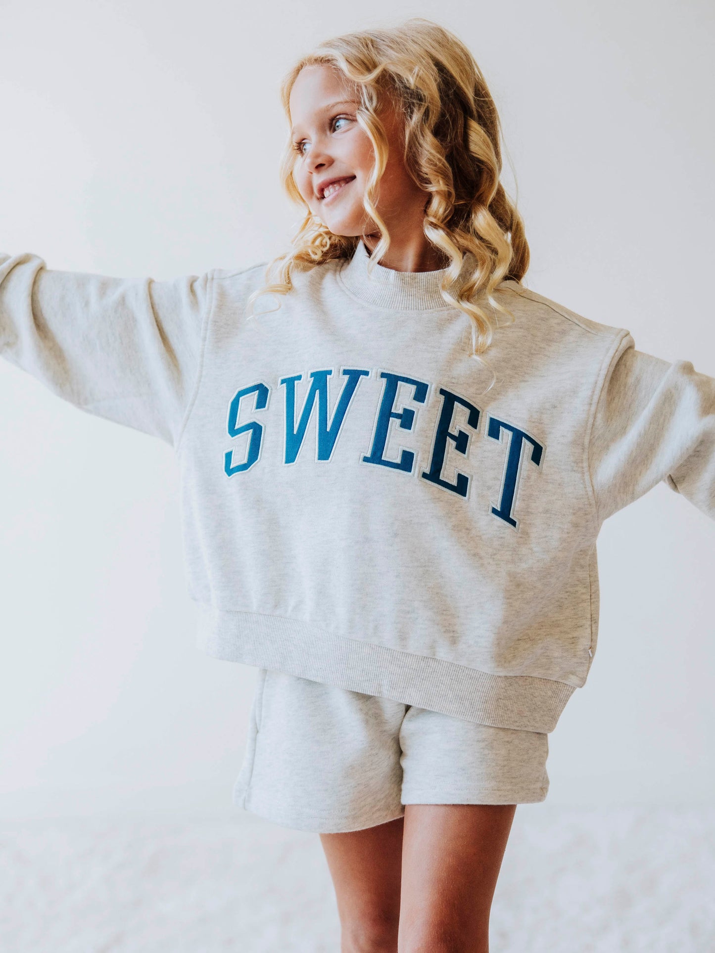 Girl model wearing a two piece gray in color set. The top says SWEET on the front in blue letters. Model is also wearing suede strapped sandals. Bottoms of set are same gray as top. 