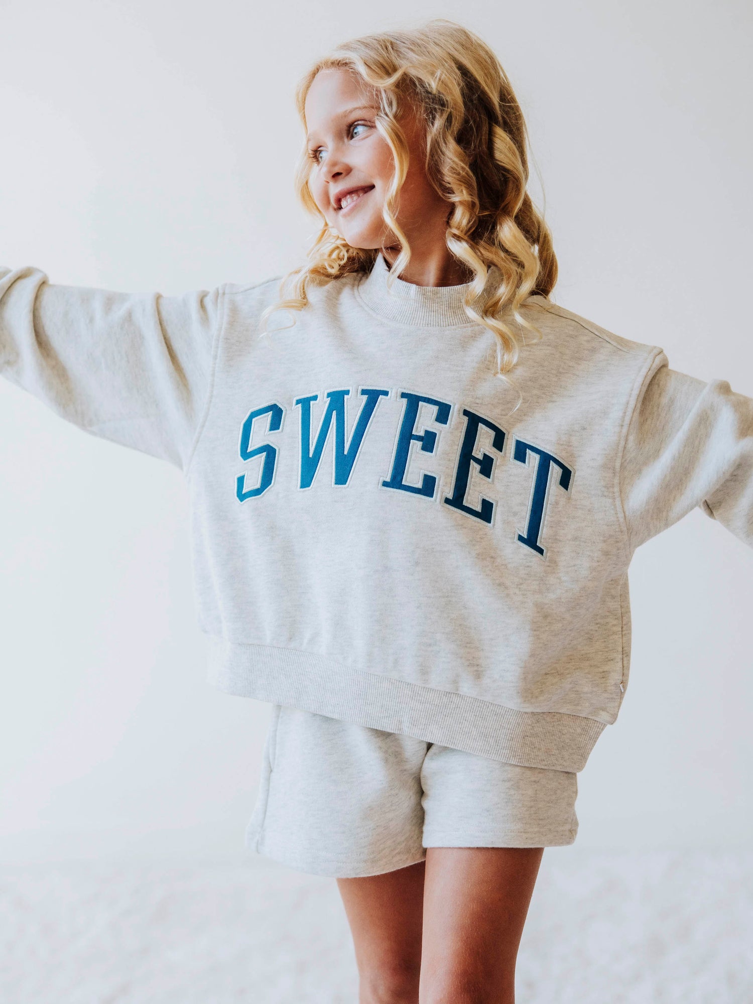Girl model wearing a two piece gray in color set. The top says SWEET on the front in blue letters. Model is also wearing suede strapped sandals. Bottoms of set are same gray as top. 