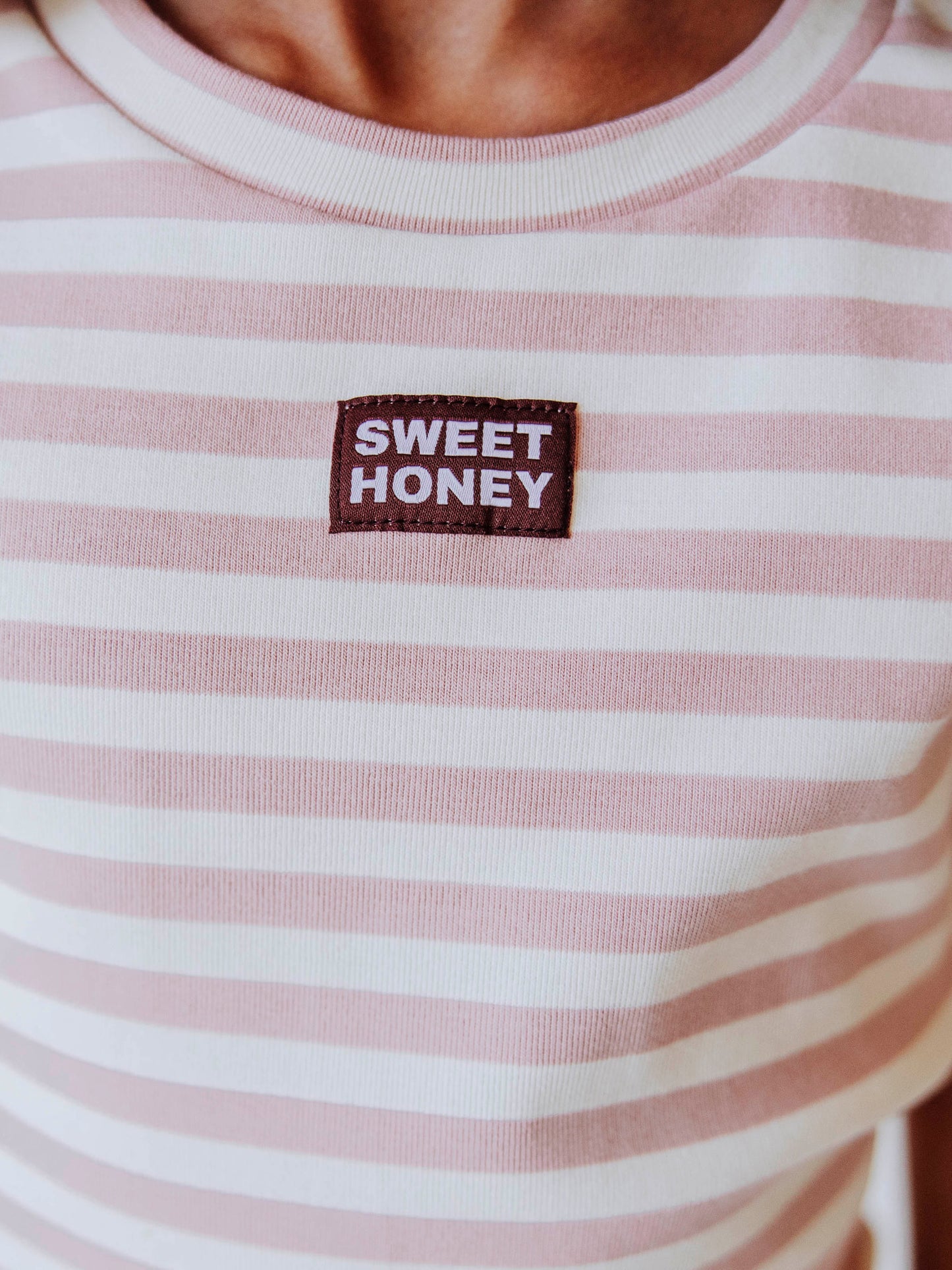 Up close shot of stitched on SweetHoney tag. Tag is brown and white. Overall shirt color is pink and ivory striped. 