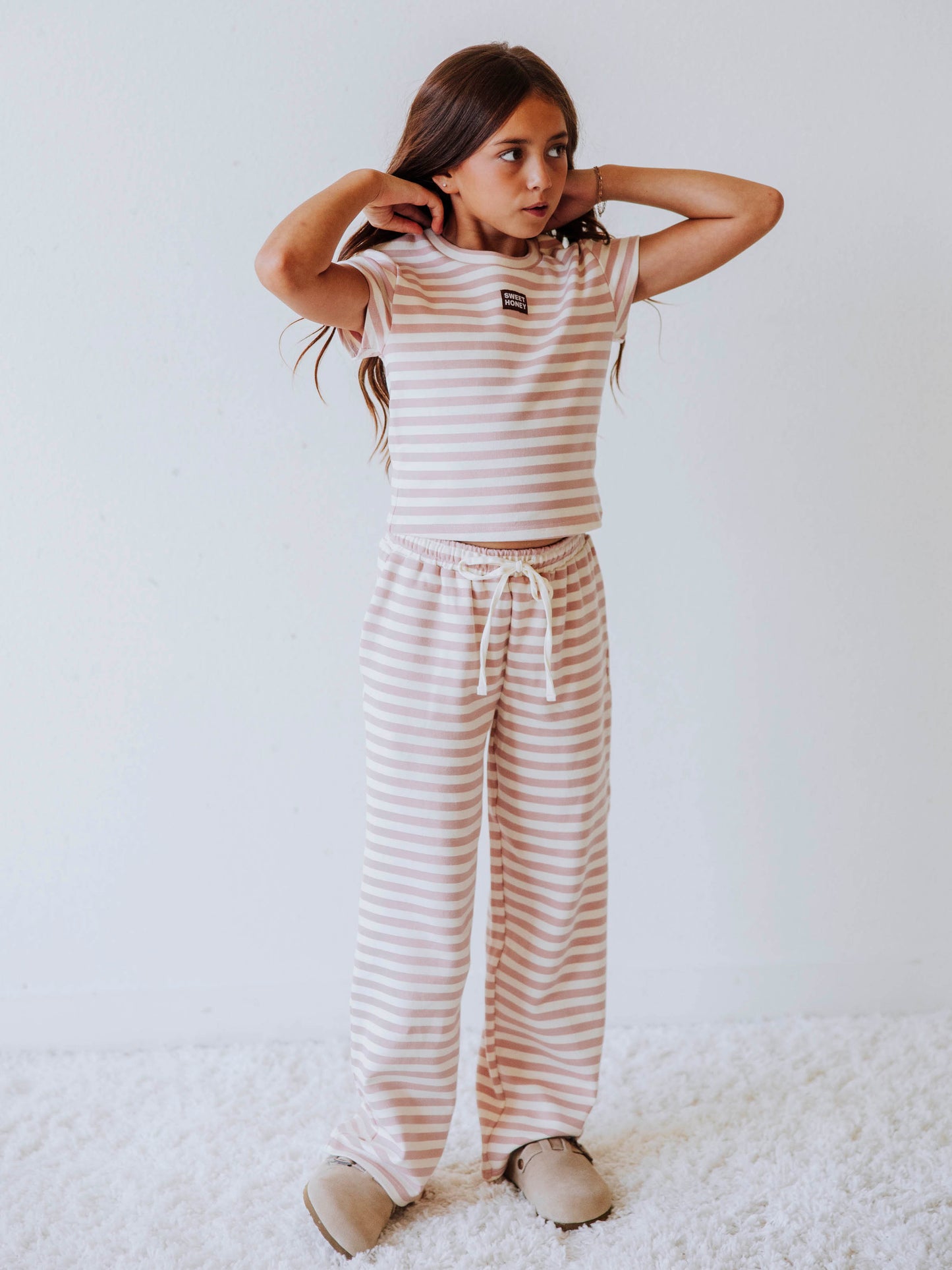 Girl is wearing a two-piece, pink and ivory striped pant set. There is a SweetHoney logo tag stitched on the center of shirt. Model is wearing clogs. 
