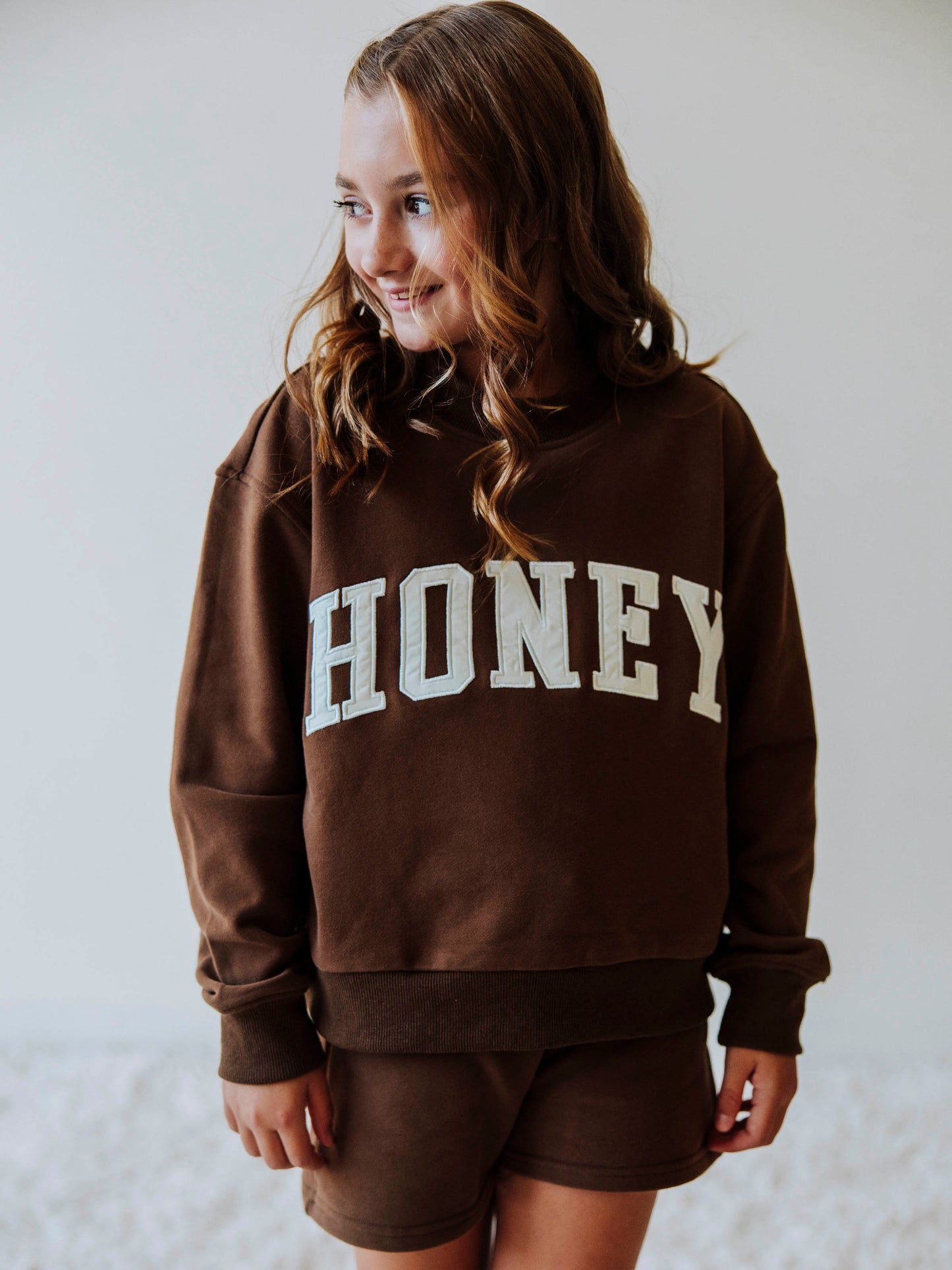 Model is wearing a two-piece, brown colored mock neck style set. Top has HONEY across the top in ivory colored letters. Bottoms are the same color as top. 