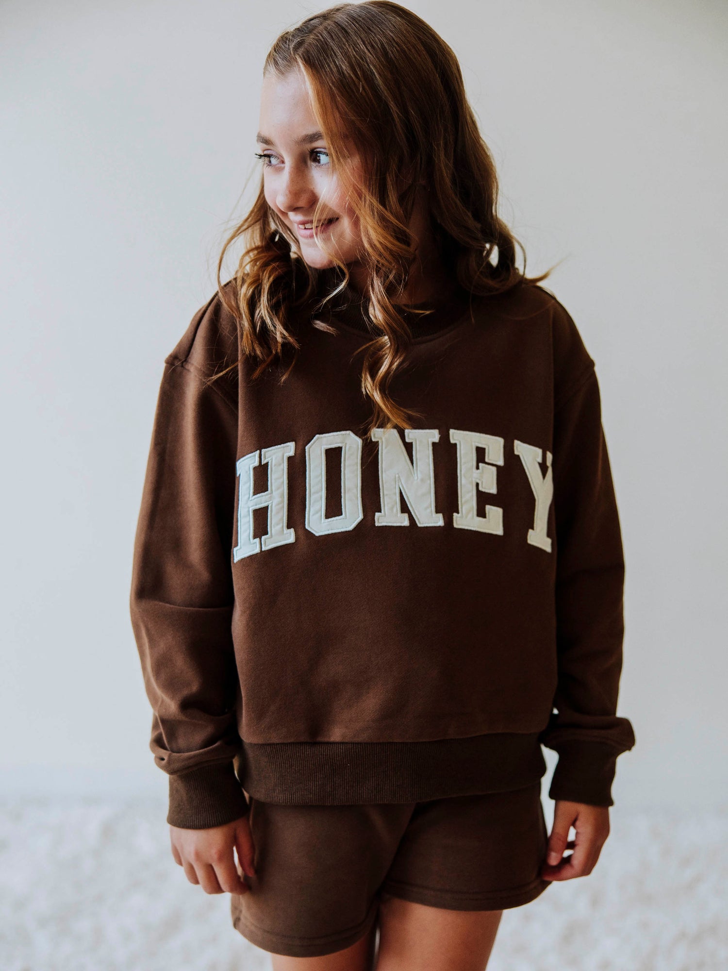 Model is wearing a two-piece, brown colored mock neck style set. Top has HONEY across the top in ivory colored letters. Bottoms are the same color as top. 