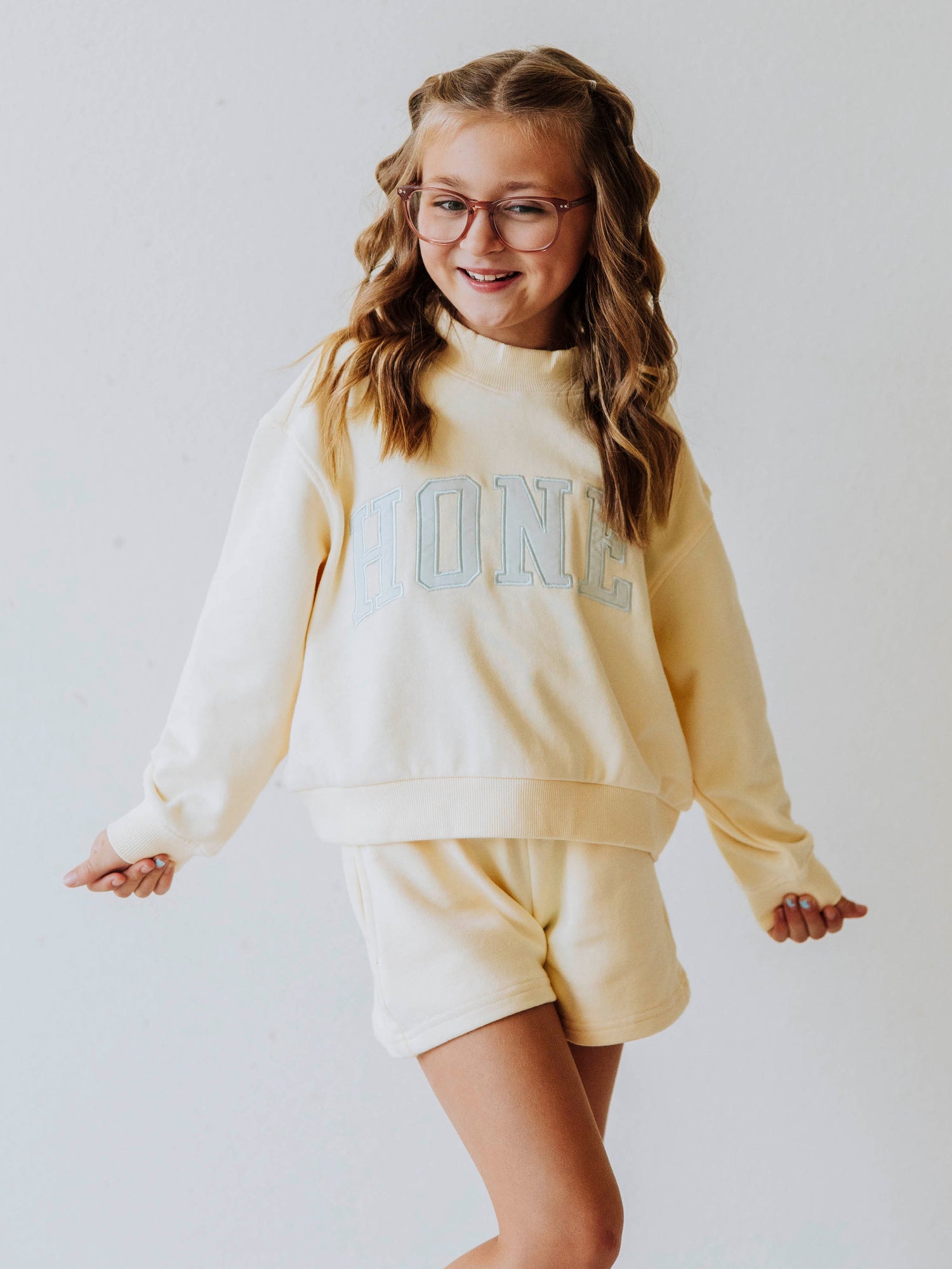 Model wearing a two piece yellow mock neck set. Color is soft. The top has the word HONEY on it in light blue. Bottoms to the set are the same color as top. 