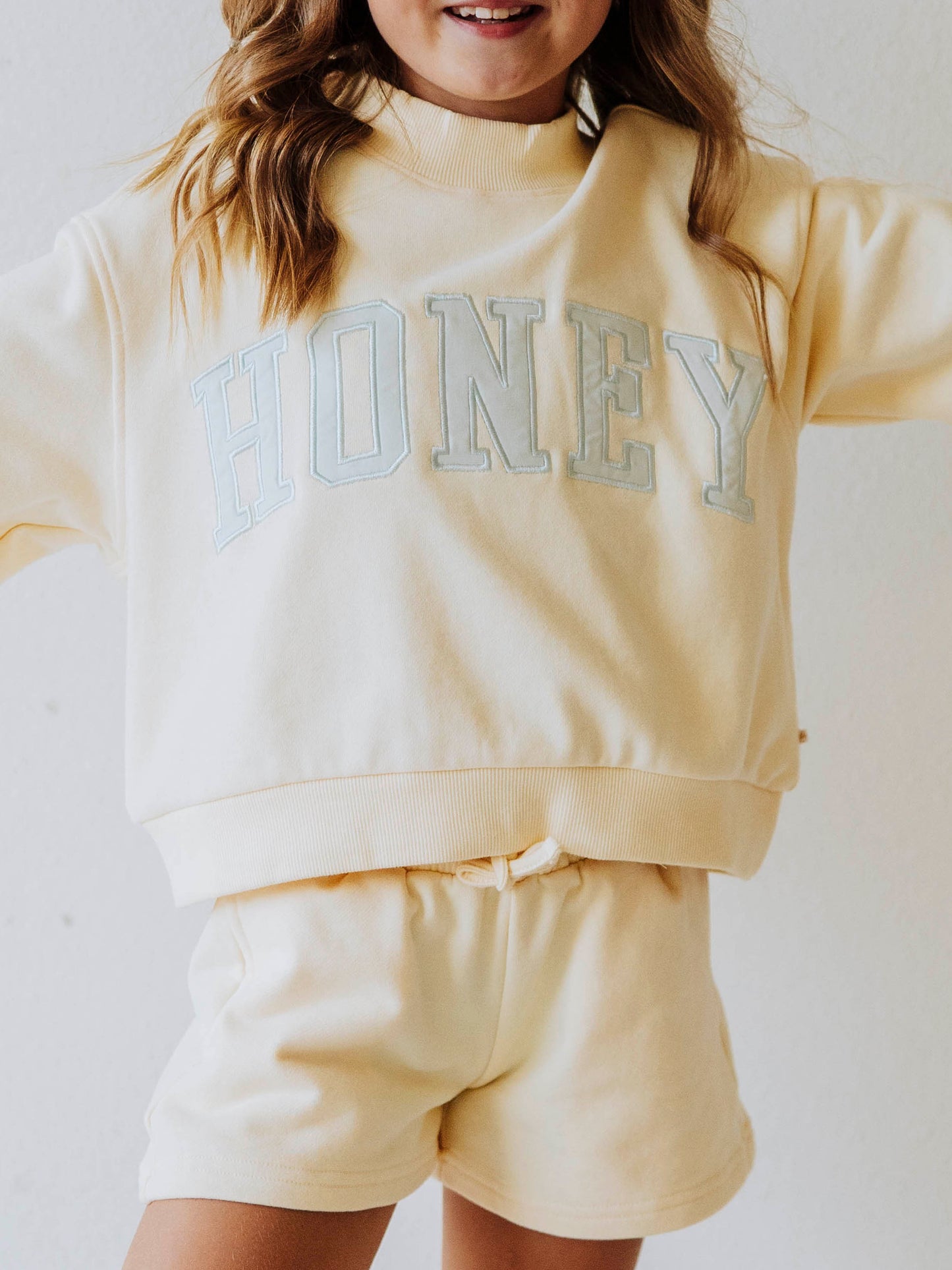 Model wearing a two piece yellow mock neck set. Color is soft. The top has the word HONEY on it in light blue. Bottoms to the set are the same color as top. 