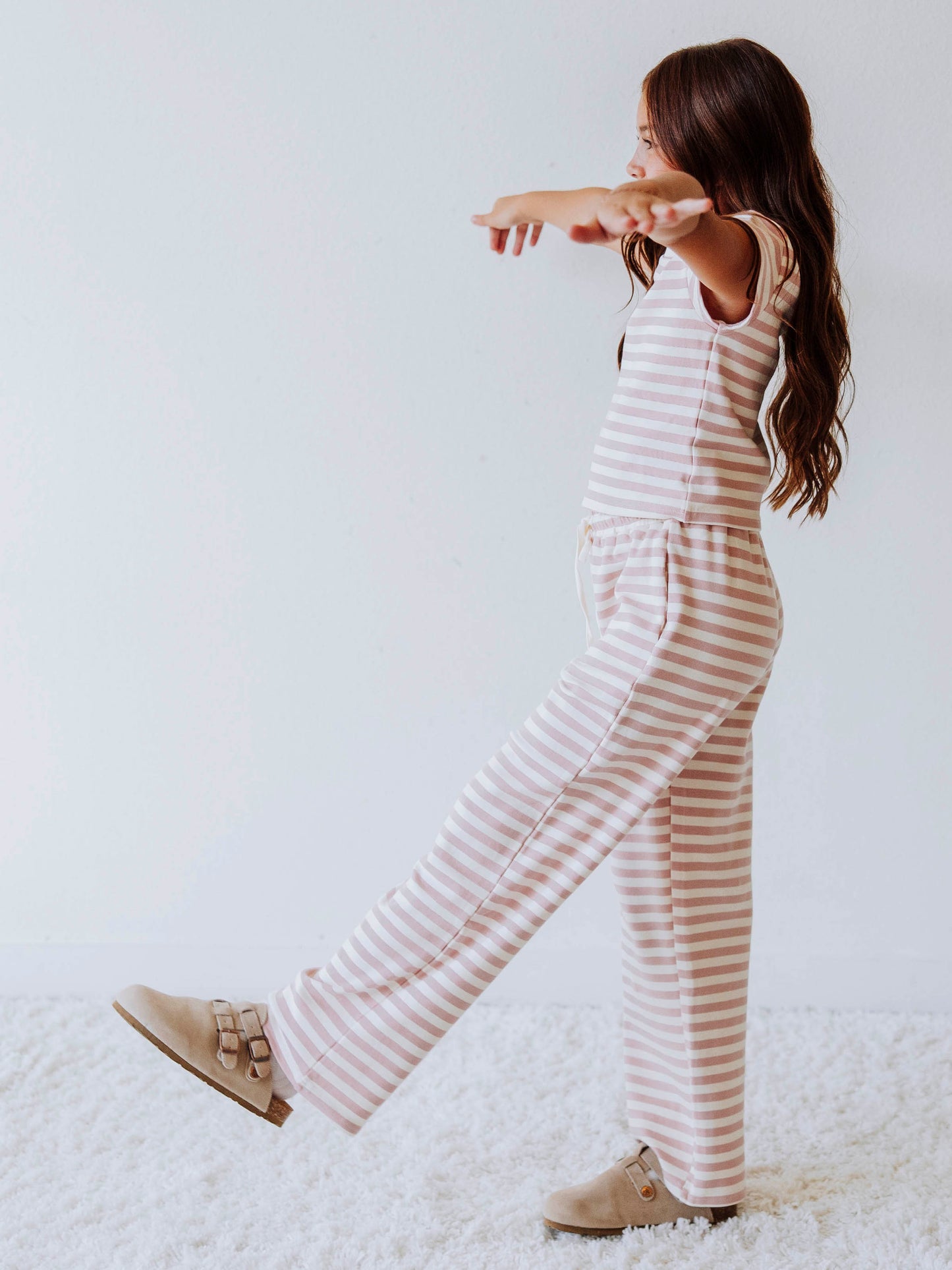 Girl is wearing a two-piece, pink and ivory striped pant set. There is a SweetHoney logo tag stitched on the center of shirt. Model is wearing clogs. 