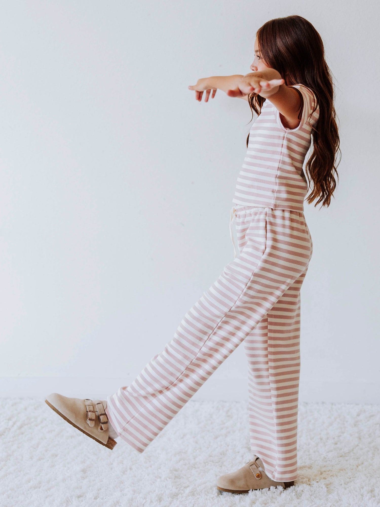 Girl is wearing a two-piece, pink and ivory striped pant set. There is a SweetHoney logo tag stitched on the center of shirt. Model is wearing clogs. 