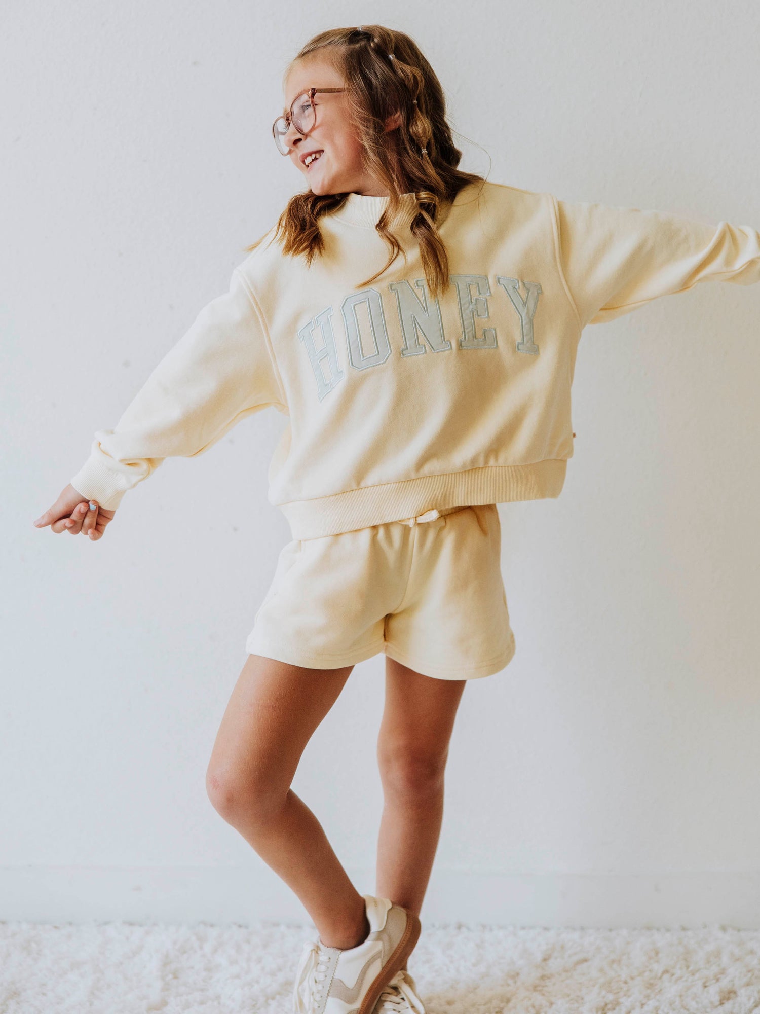 Model wearing a two piece yellow mock neck set. Color is soft. The top has the word HONEY on it in light blue. Bottoms to the set are the same color as top. 
