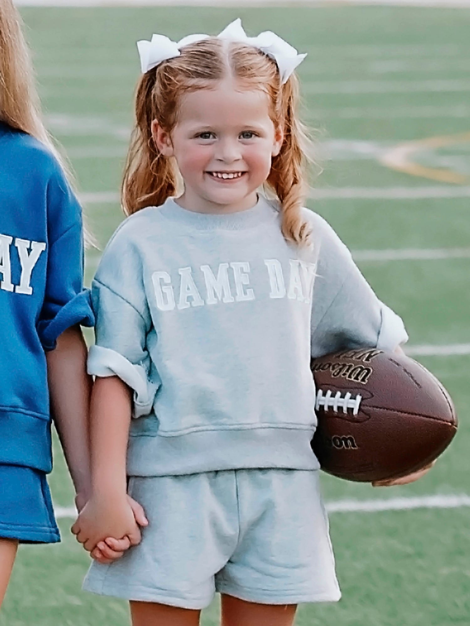 Girl outside wearing a two-piece gray sweat set that says GAME DAY on the front. Girl is holding a football.