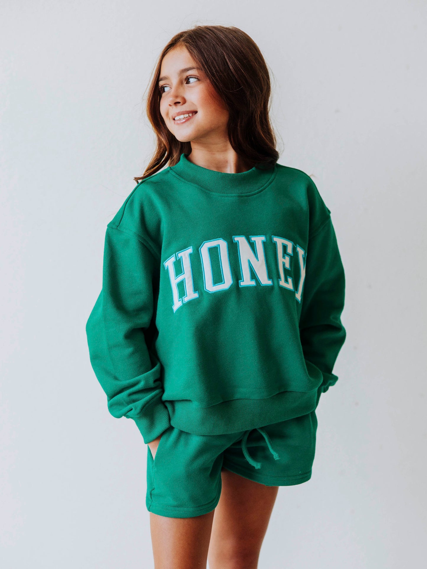 Model is wearing a two-piece, teal mock neck set, with HONEY on across the top of the top. Shorts are the same color as top. 