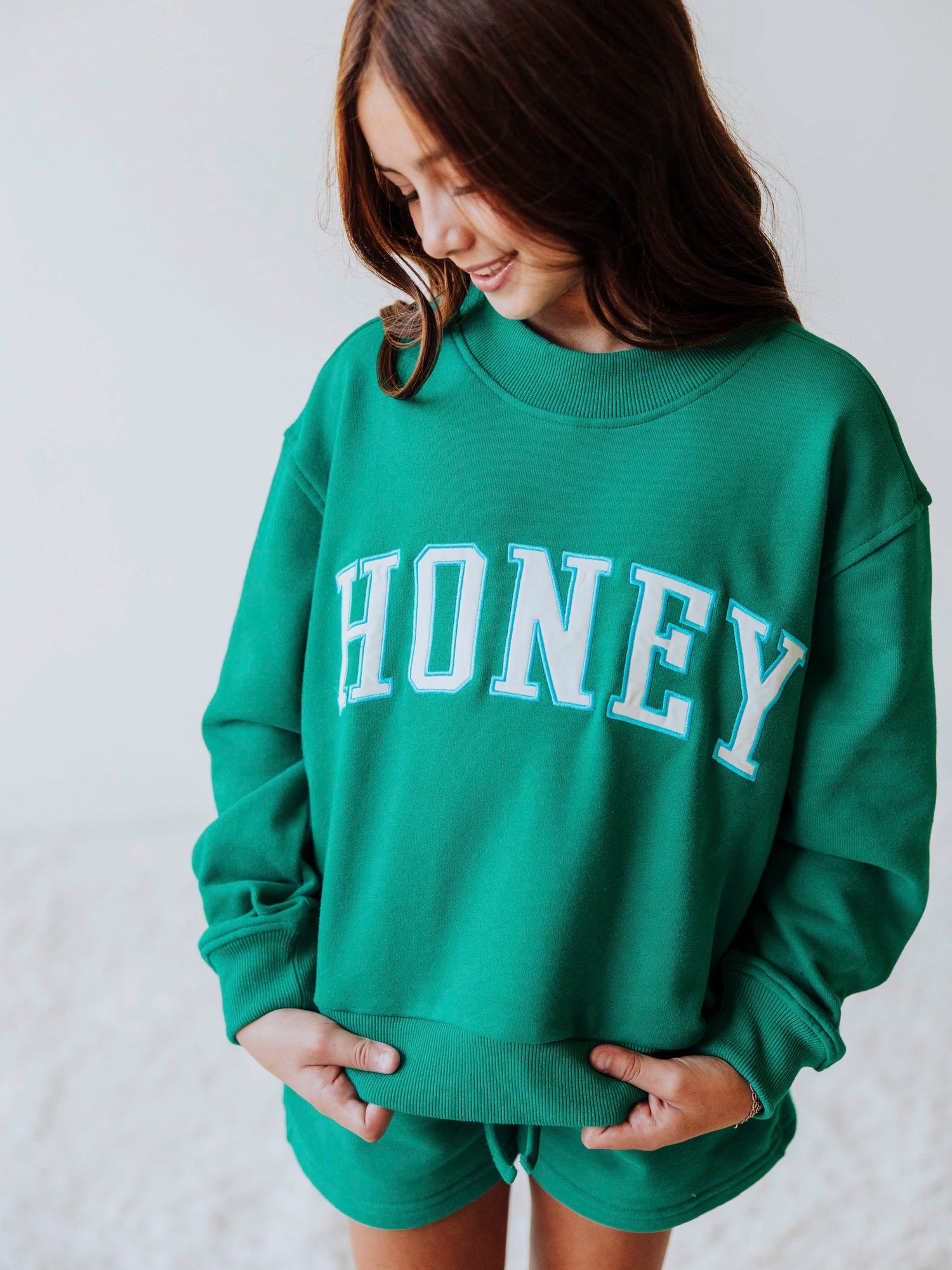 Model is wearing a two-piece, teal mock neck set, with HONEY on across the top of the top. Shorts are the same color as top. 