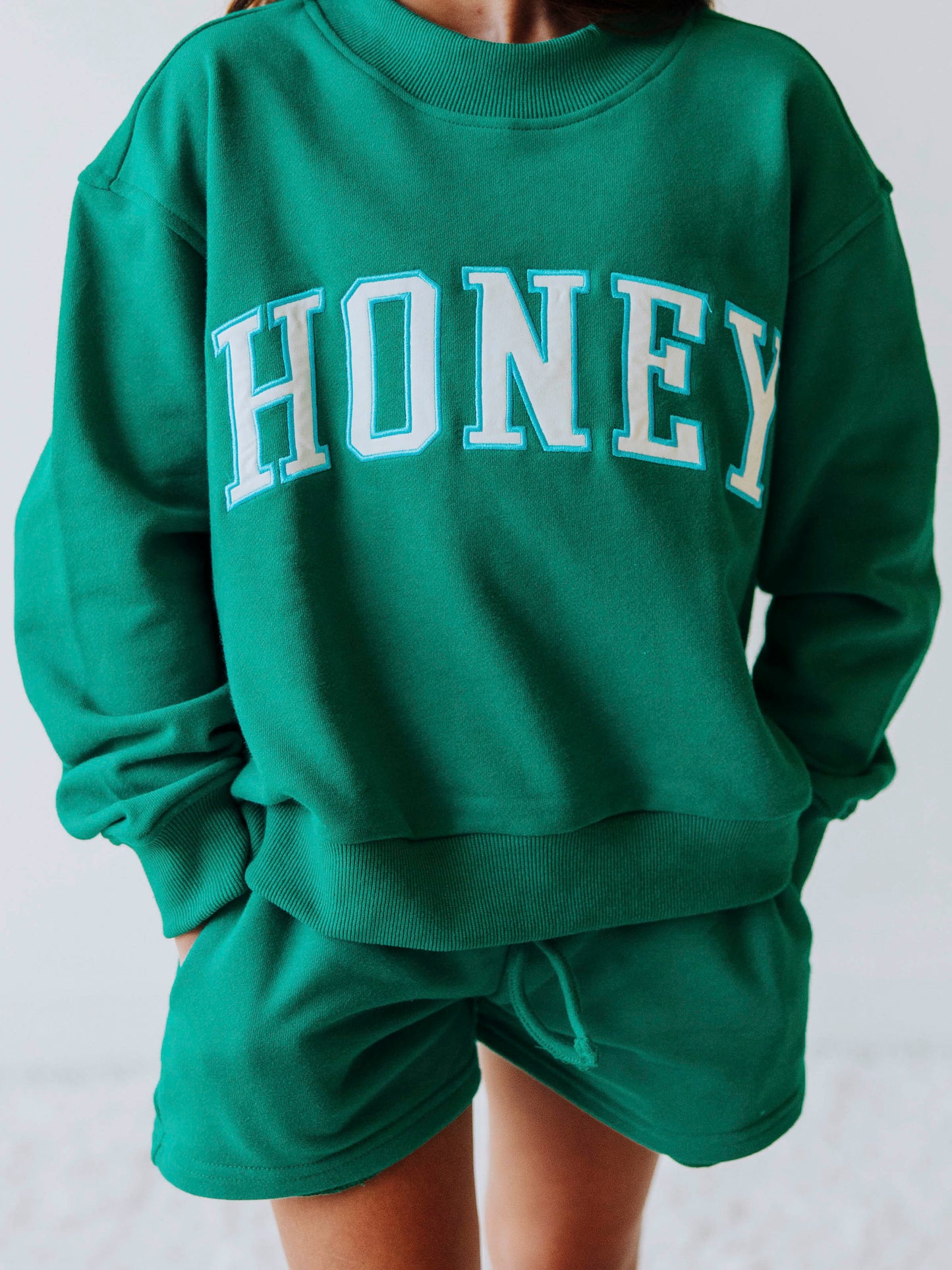 Model is wearing a two-piece, teal mock neck set, with HONEY on across the top of the top. Shorts are the same color as top. 