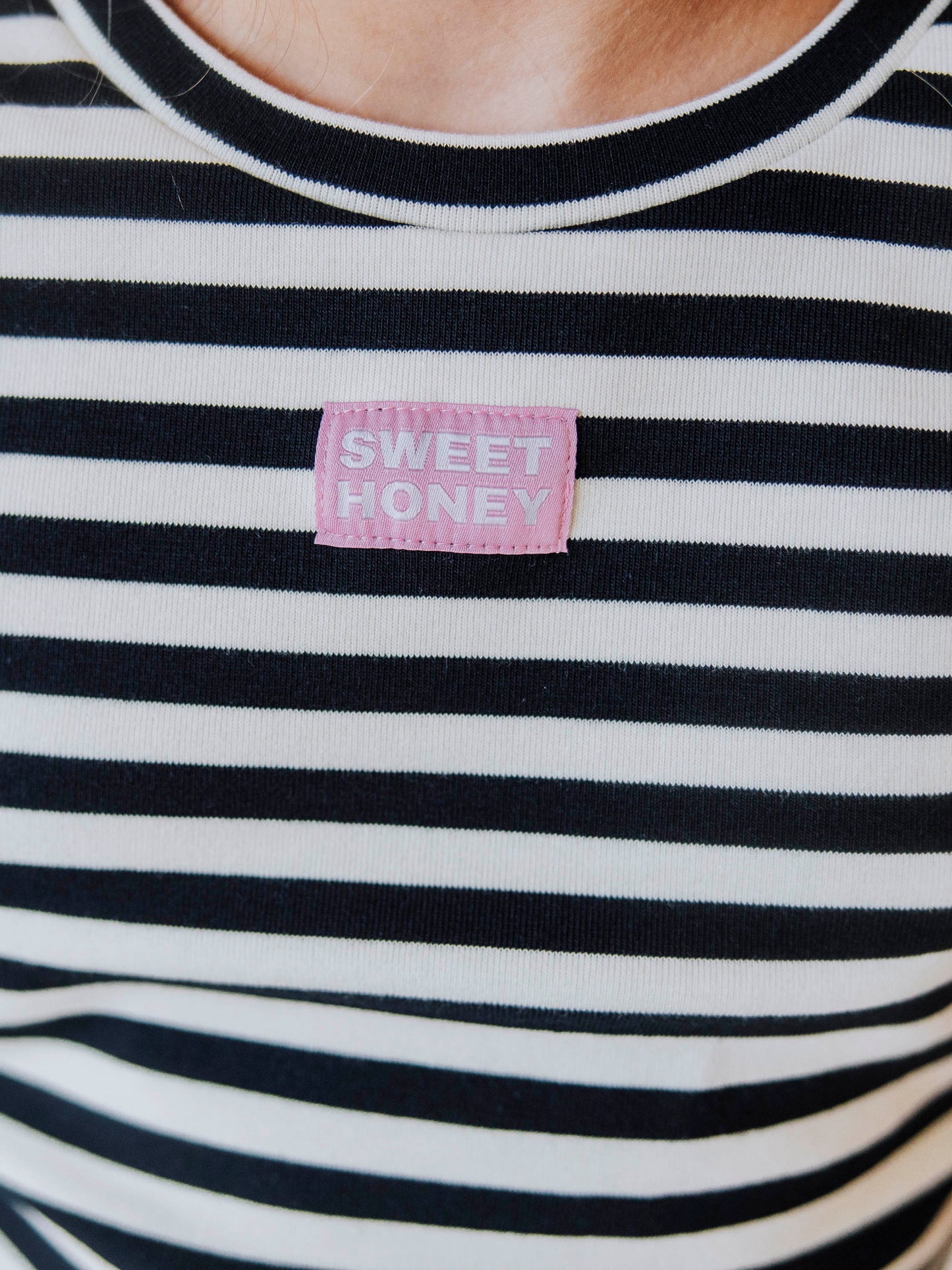 Girl is wearing a two-piece, navy and ivory striped pant set. There is a SweetHoney logo tag stitched on the center of shirt. This shot is up close of the tag. 