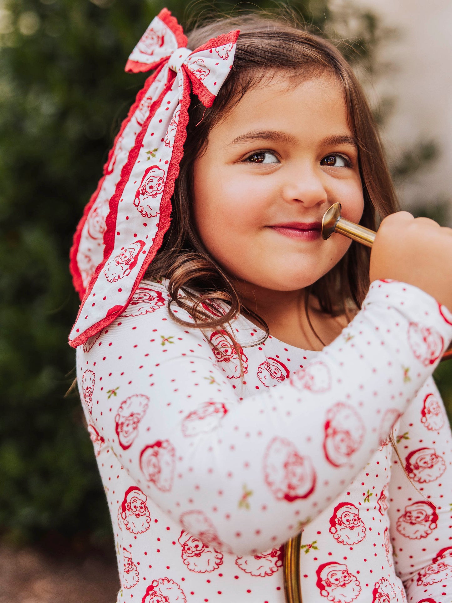Girl wearing a long tail santa bow with polka dots. Bow is trimmed in red lace. Model's outfit is the same print as bow. Model has a mouth piece to a trumpet to her mouth