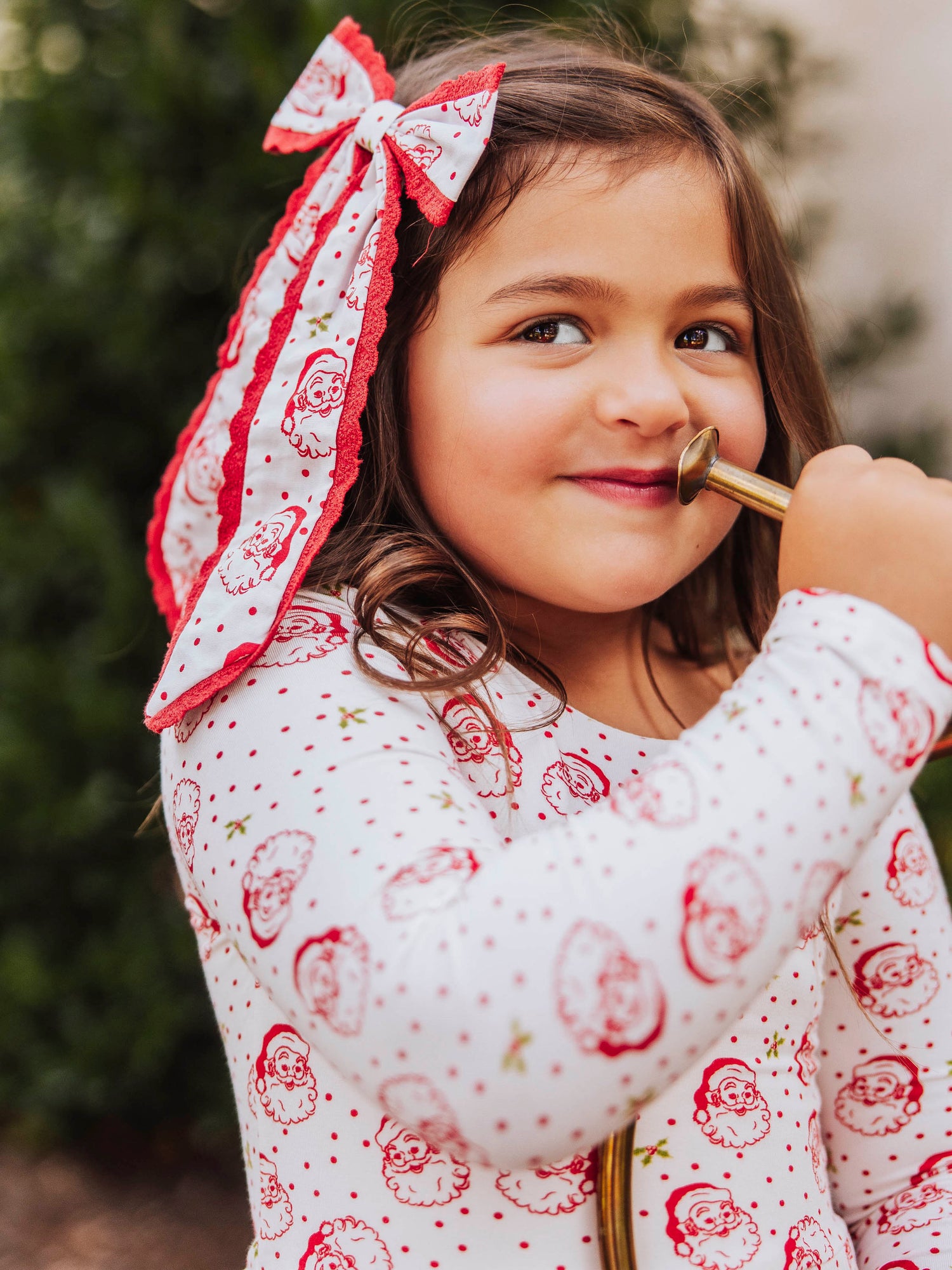 Girl wearing a long tail santa bow with polka dots. Bow is trimmed in red lace. Model's outfit is the same print as bow. Model has a mouth piece to a trumpet to her mouth