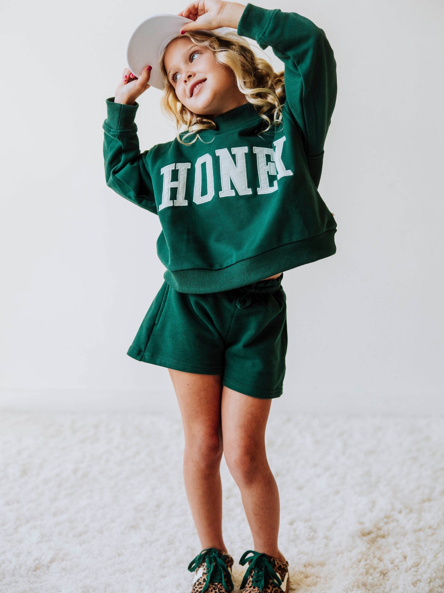 model wearing a two-piece, dark green, mock neck set that says HONEY on the front in a white/ivory color. Model is also wearing leopard shoes with green laces and is wearing a white hat. 