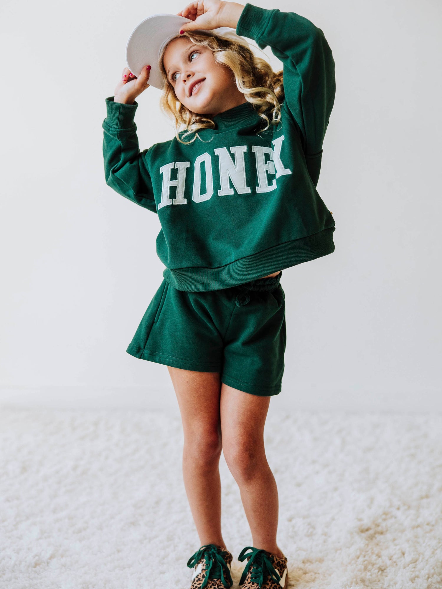 model wearing a two-piece, dark green, mock neck set that says HONEY on the front in a white/ivory color. Model is also wearing leopard shoes with green laces and is wearing a white hat. 