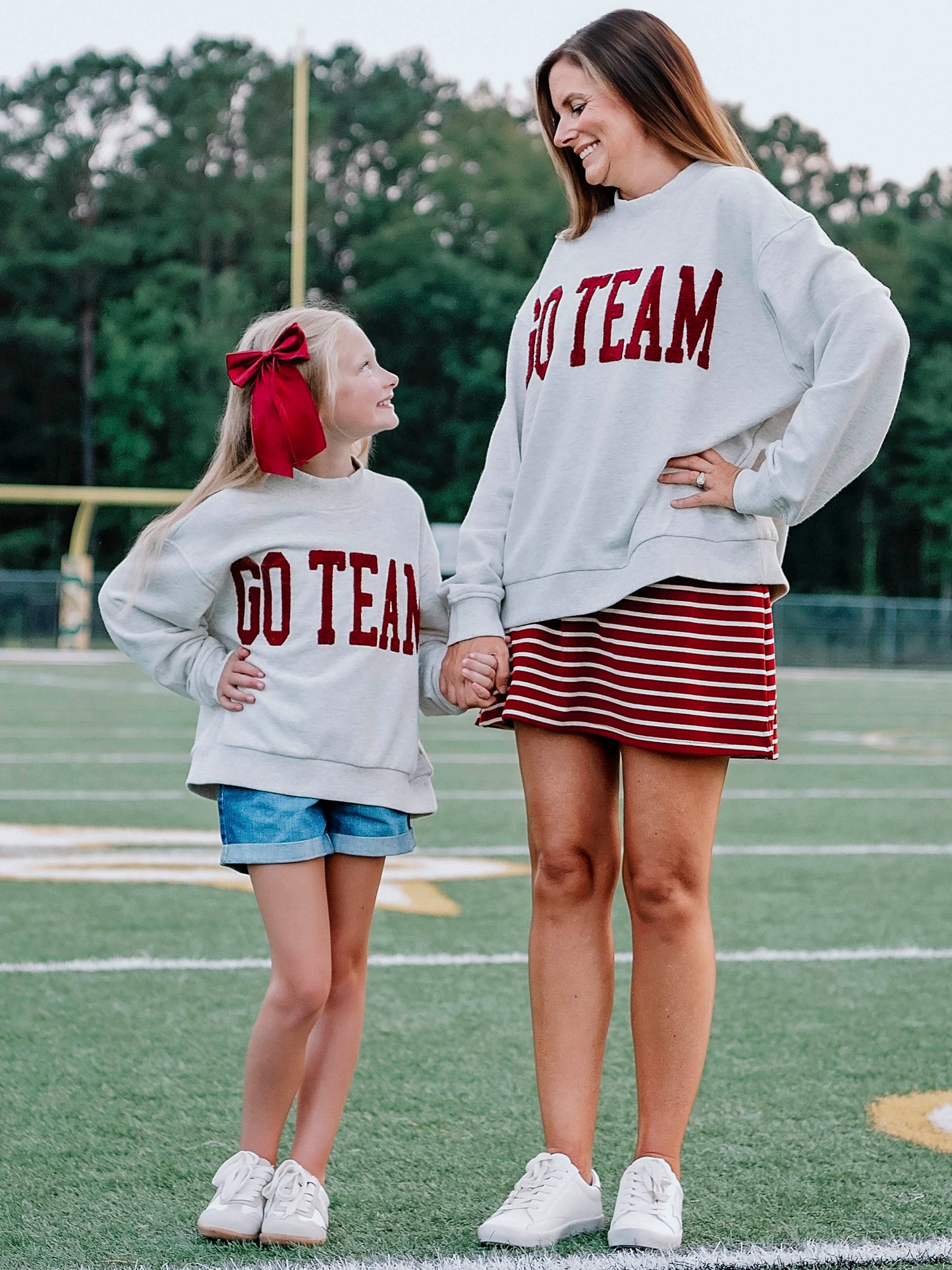 Women outside wearing a heathered gray sweater that says GO TEAM in a crimson red color. She is holding hands with a small child wearing the same sweater. 