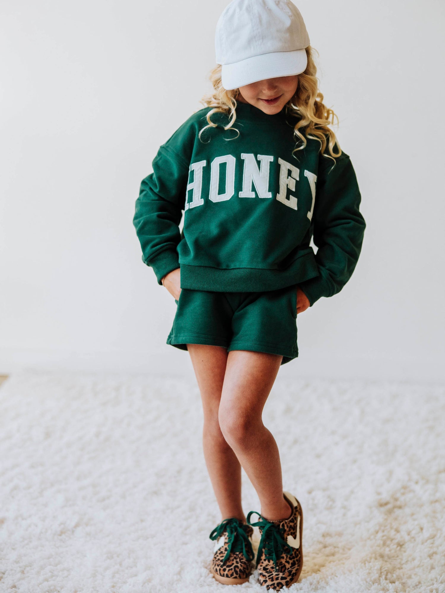 model wearing a two-piece, dark green, mock neck set that says HONEY on the front in a white/ivory color. Model is also wearing leopard shoes with green laces and is wearing a white hat. 