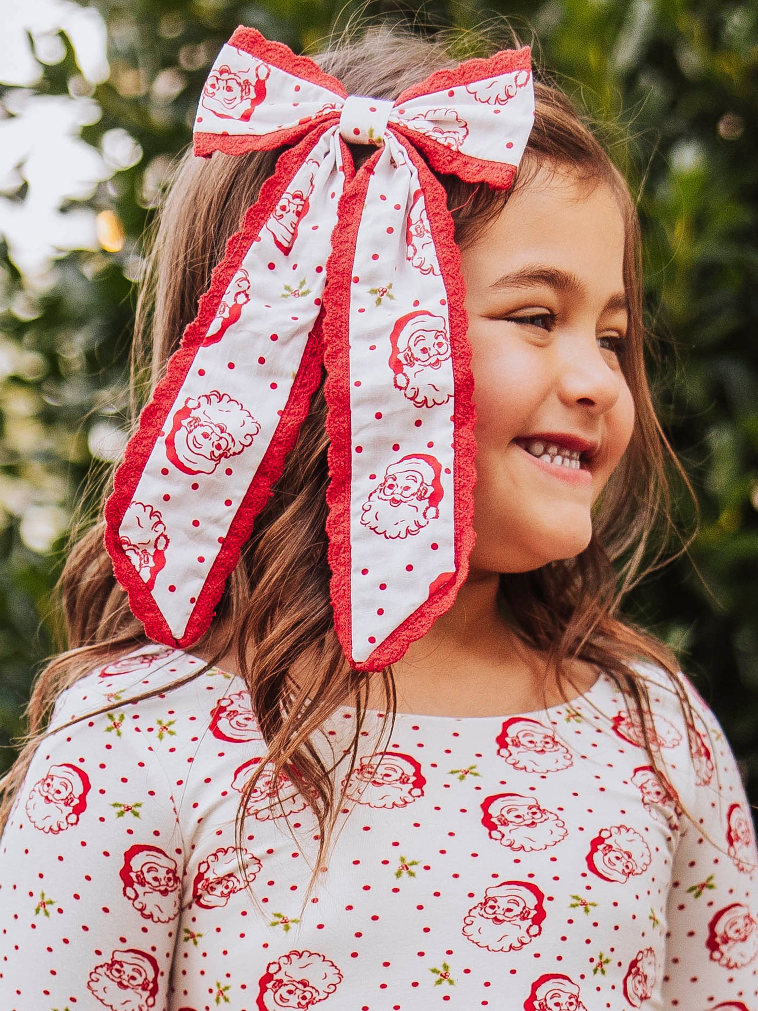 Girl wearing a long tail santa bow with polka dots. Bow is trimmed in red lace. Model's outfit is the same print as bow. 