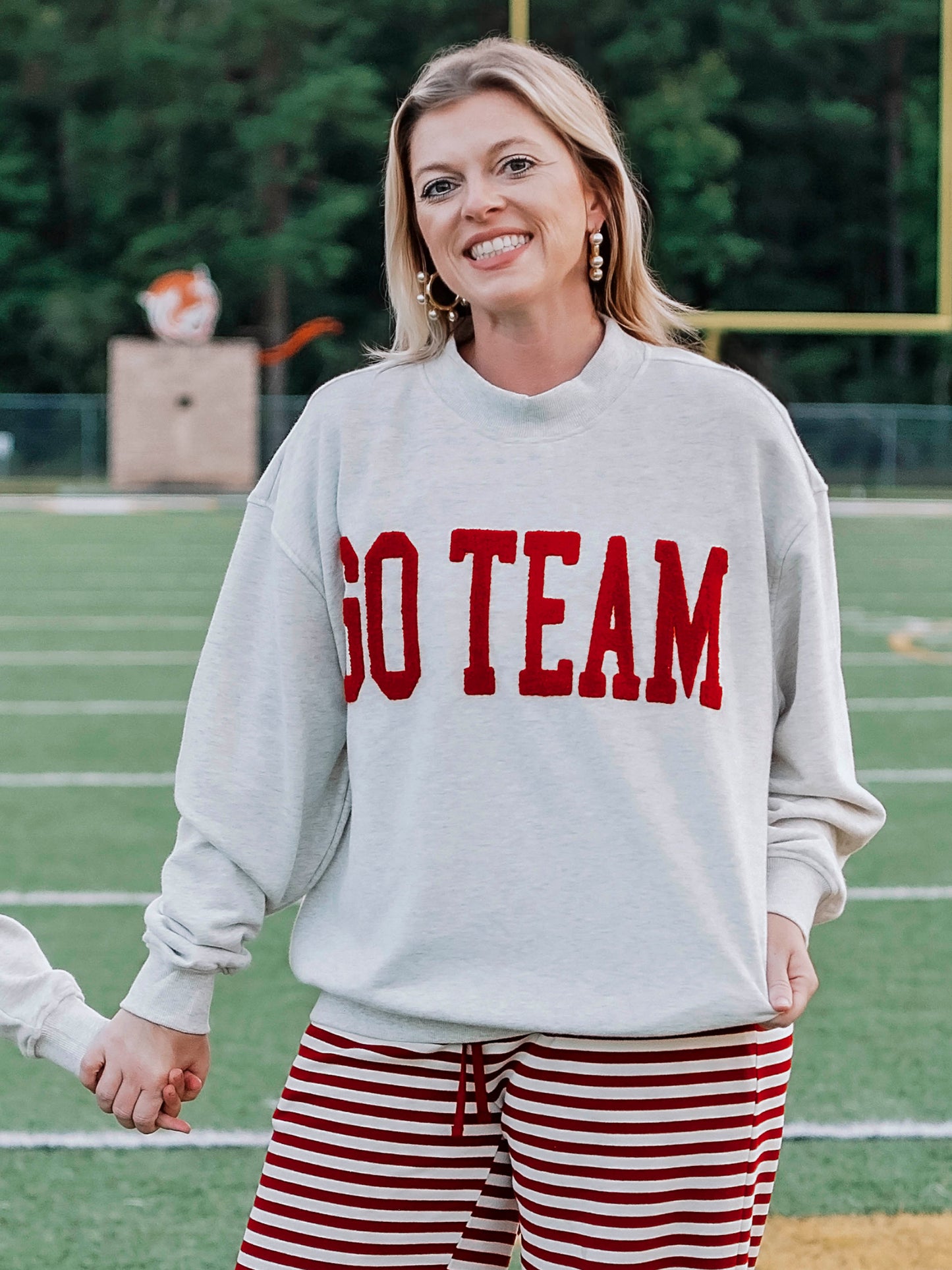 Women outside wearing a heathered gray sweatshirt that says GO TEAM in a crimson red color. She is holding hands with someone. 