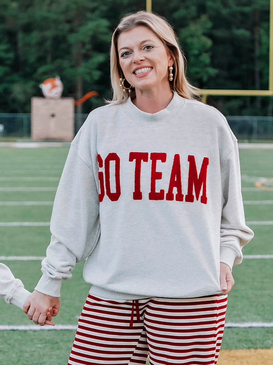 Women outside wearing a heathered gray sweatshirt that says GO TEAM in a crimson red color. She is holding hands with someone. 