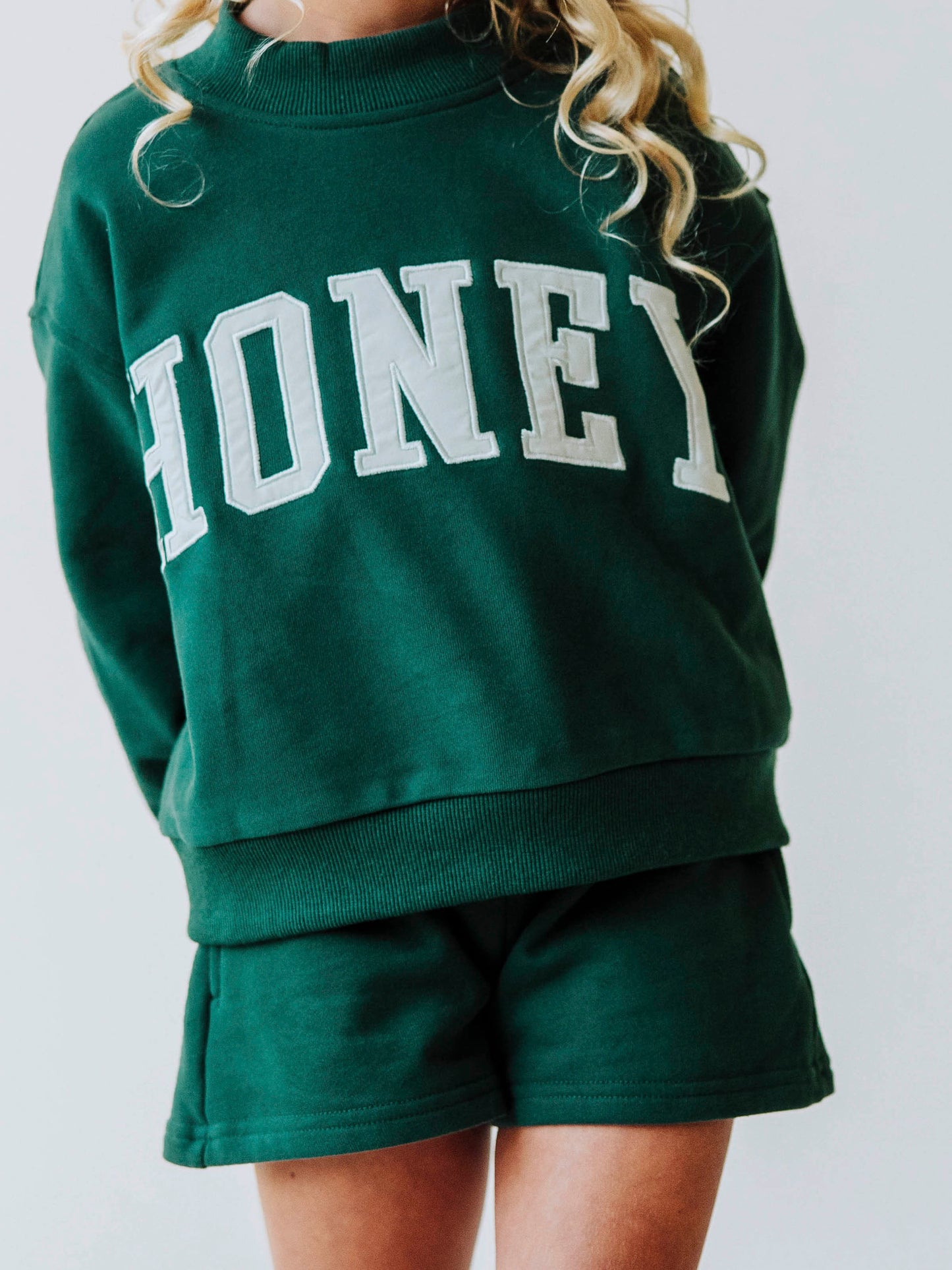 Close up of model wearing a two-piece, dark green colored mock neck style set. Top has HONEY across the top in ivory colored letters. Shorts are the same color as the top. 
