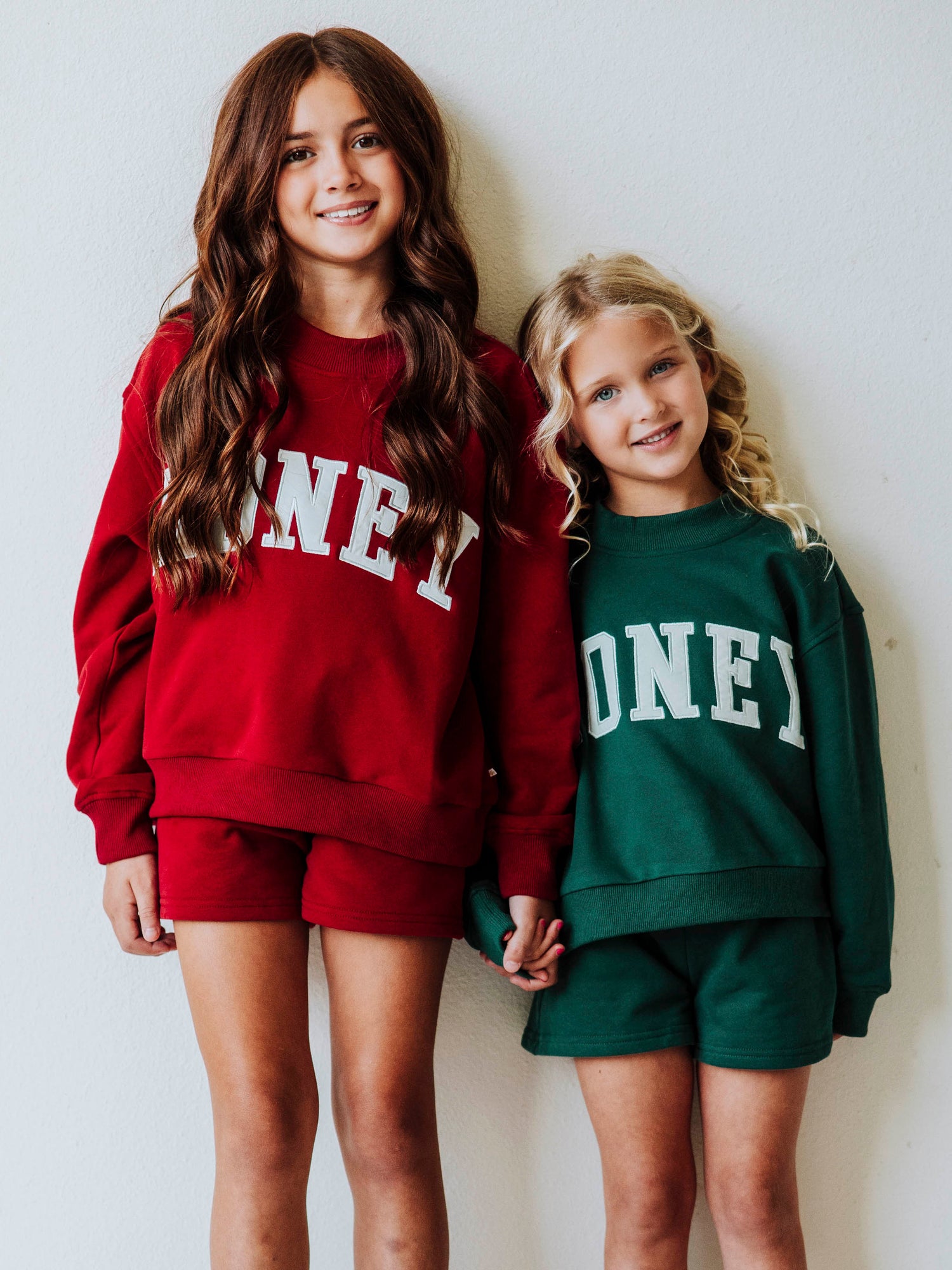 Picture is of two girl models both wearing two-piece, mock neck sets. One set is crimson in color and the other is dark green. Both tops say HONEY across the front in ivory letters. 