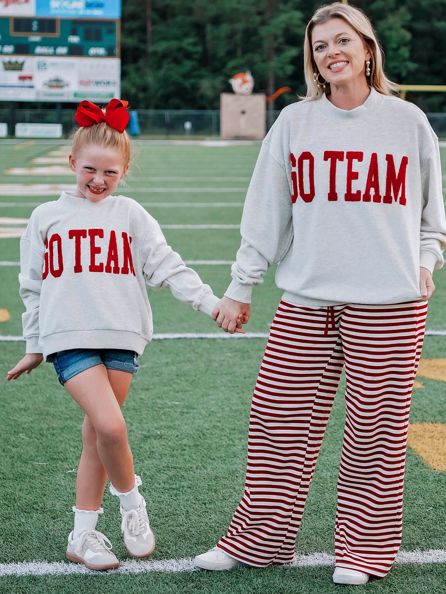 Women outside wearing a heathered gray sweatshirt that says GO TEAM in a crimson red color. She is holding hands with a little girl who is wearing the same sweater with denim shorts. 