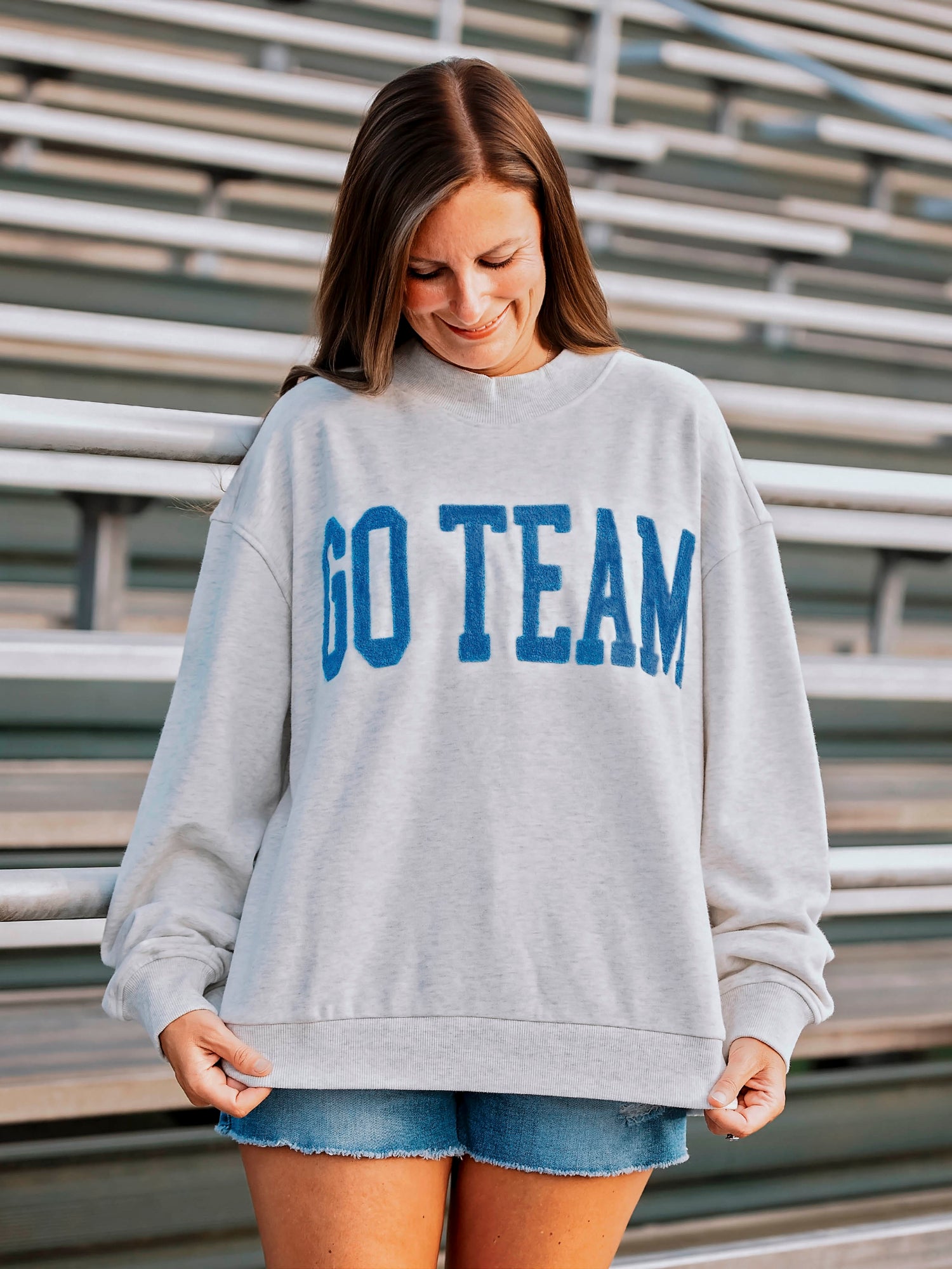 Women wearing a heathered gray sweatshirt that says GO TEAM on the front in a royal blue. Women appears to be in football stands. 