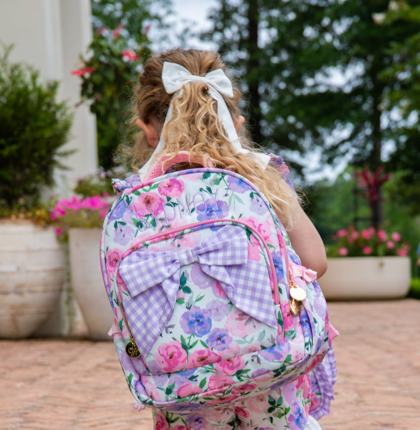 Girl wearing a colorful floral backpack with a purple gingham bow attached to the backpack. Pack also has custom embroidery. Girl also holding same printed style lunch kit