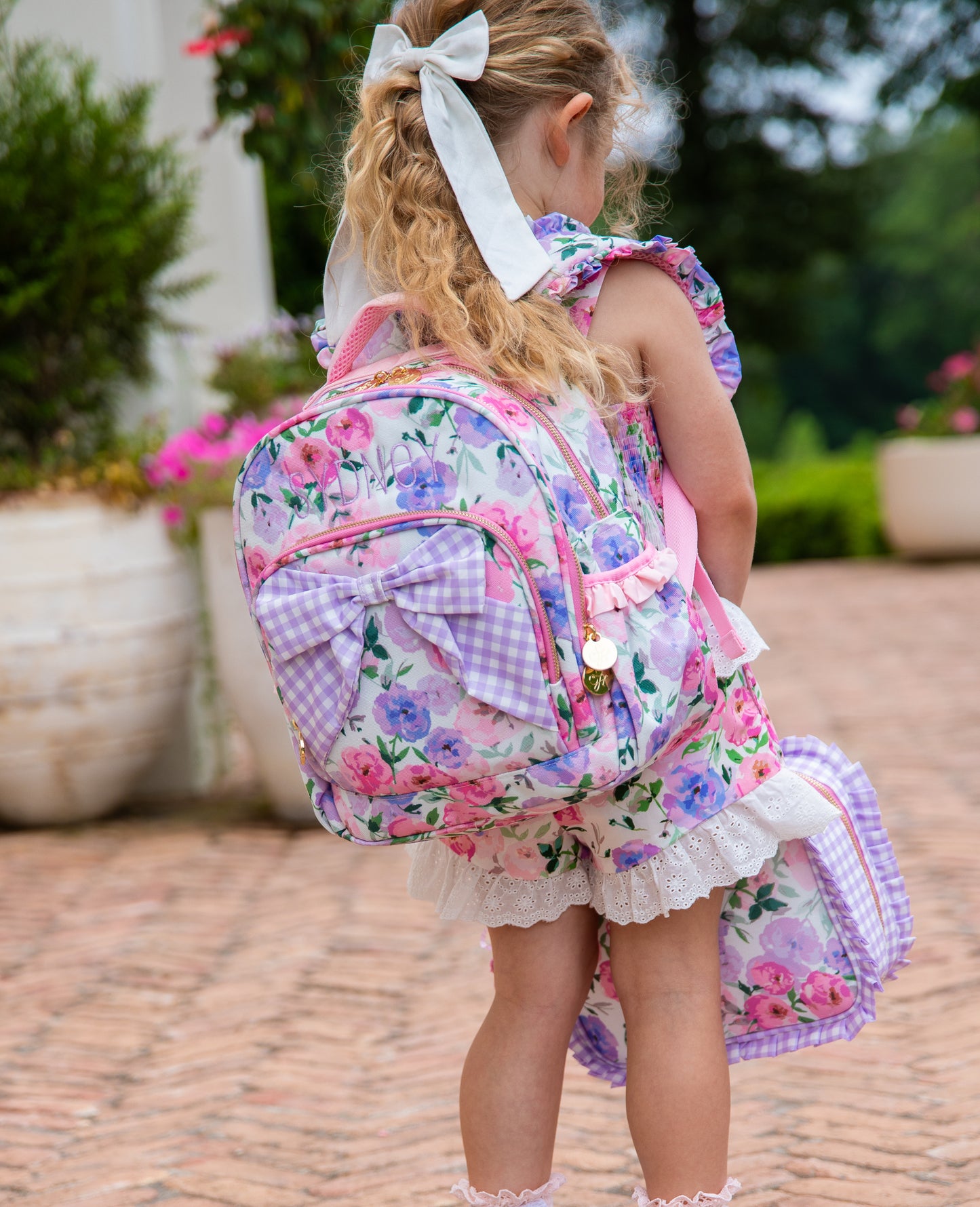 Girl wearing a colorful floral backpack with a purple gingham bow attached to the backpack. Pack also has custom embroidery. Girl also holding same printed style lunch kit