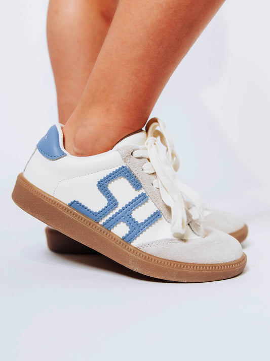Close shot of girl wearing white sneakers with "SH" in stitched blue leather on outer side of shoe and blue heel caps.