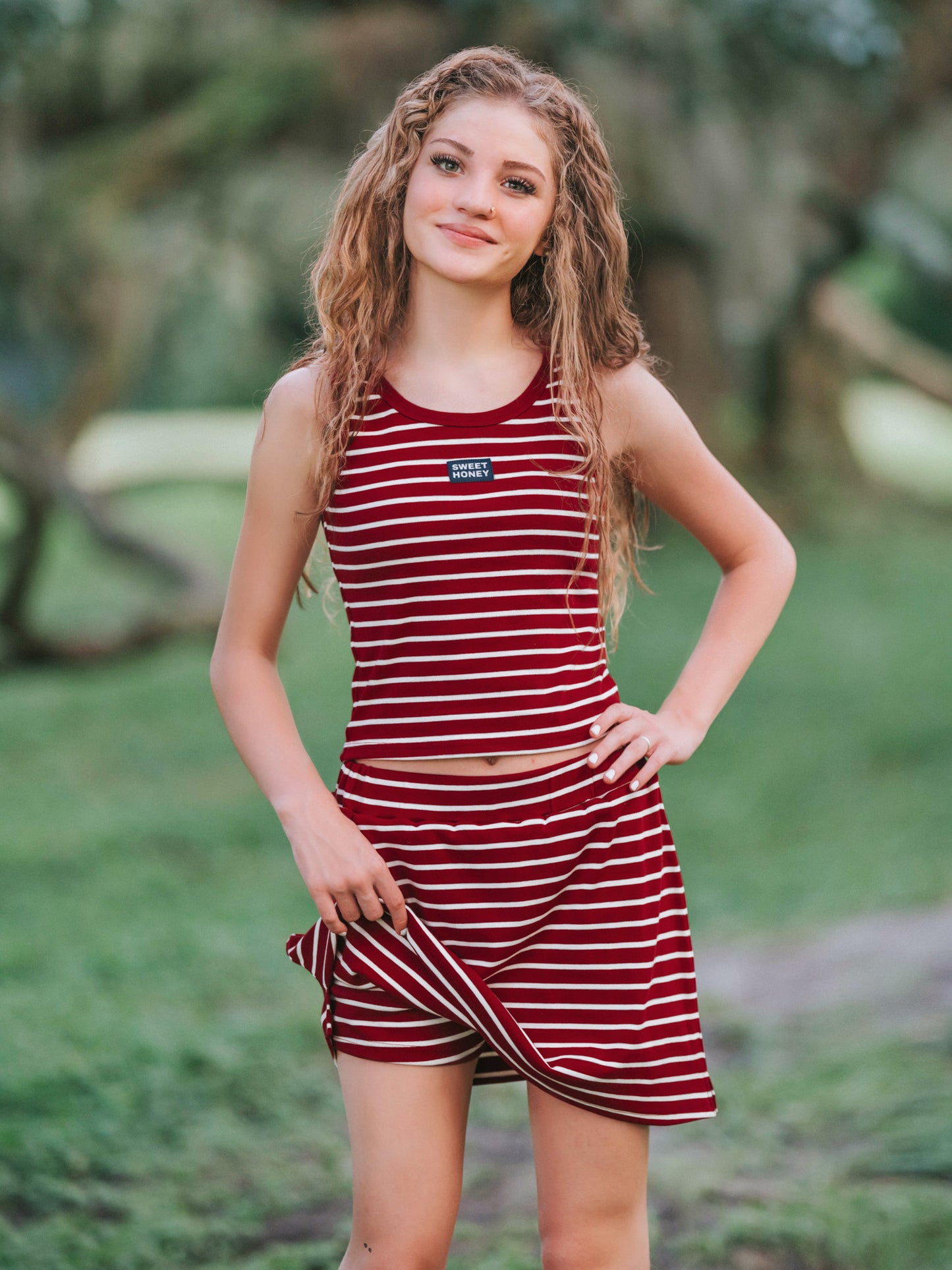 Girl outside wearing a two piece skort set that is crimson and ivory striped. Girl is slightly holding up her skort to show the built-in shorts underneath