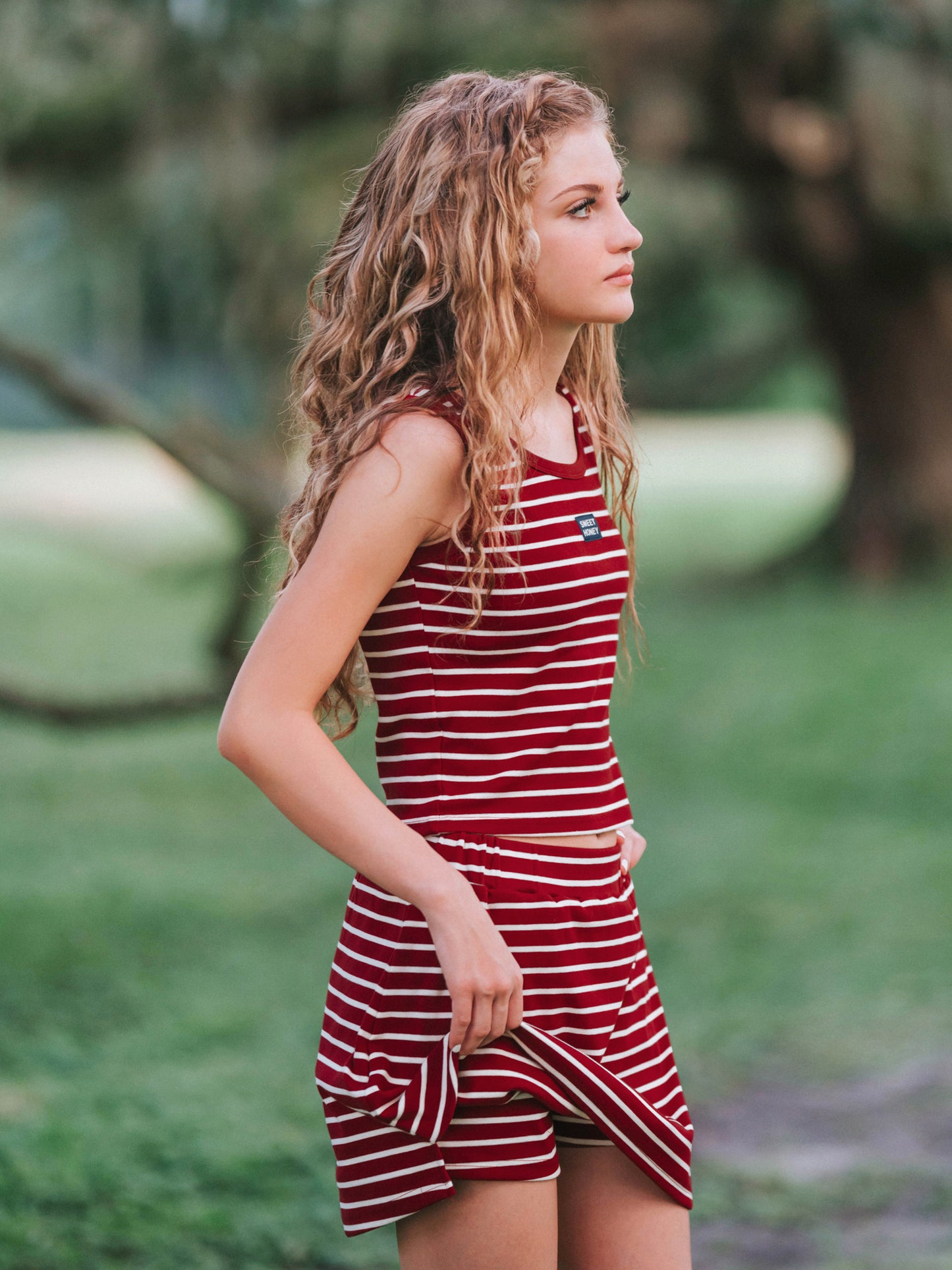 Girl outside wearing a two piece skort set that is crimson and ivory striped. Girl is slightly holding up her skort to show the built-in shorts underneath