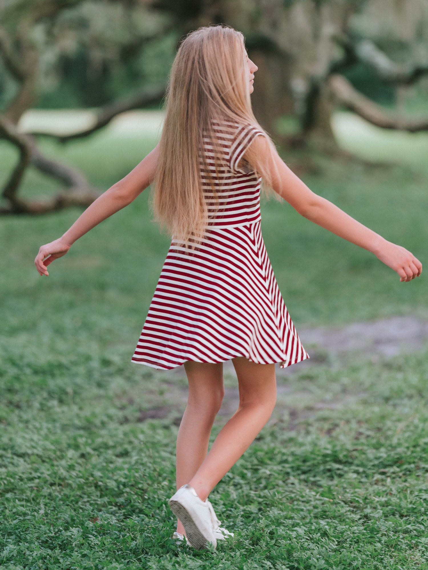 Model outside in wooded green area wearing a crimson and ivory striped dress with white canvas shoes. Model is slowly twirling.