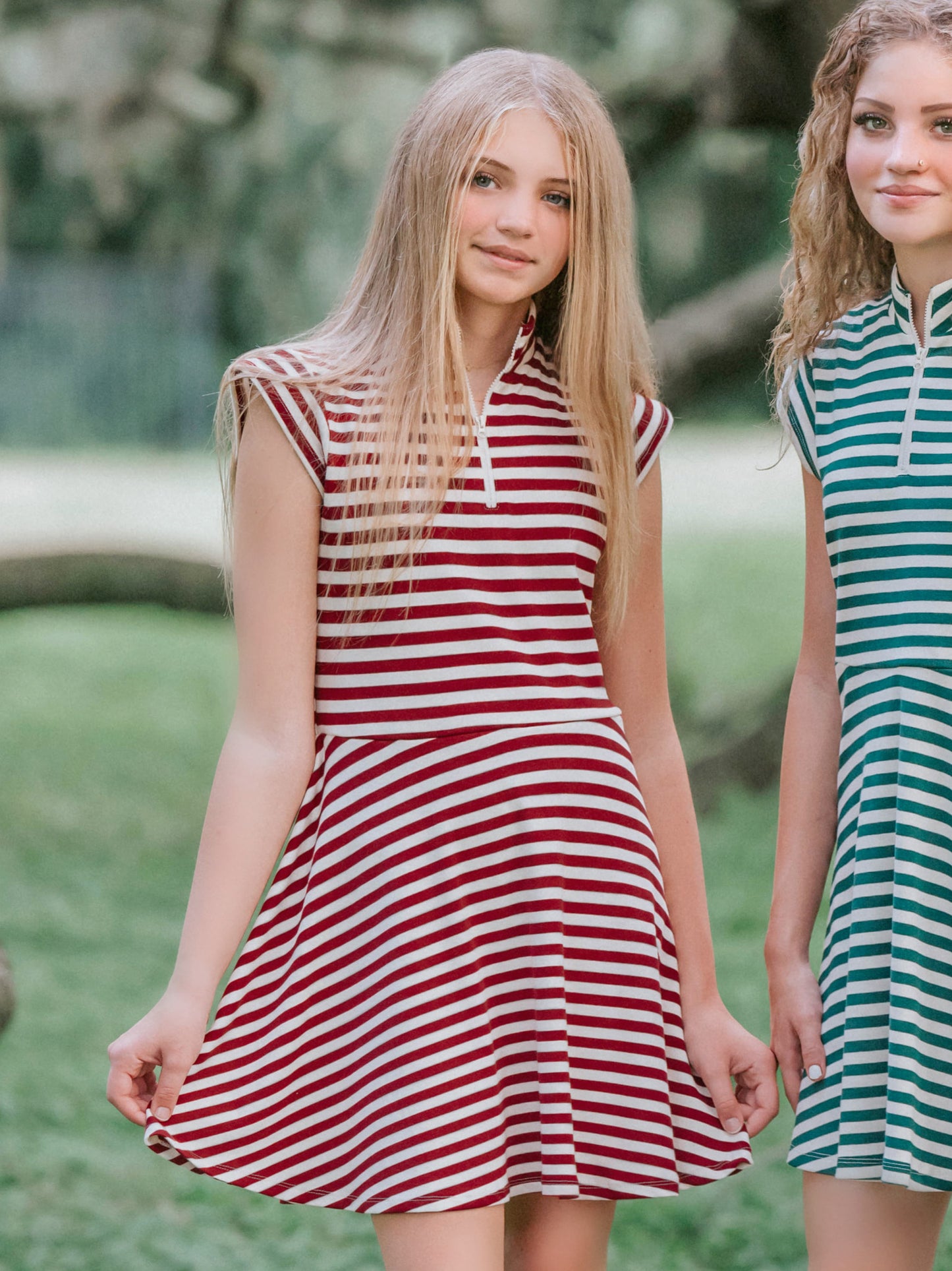 Model outside in wooded green area wearing a crimson and ivory striped dress with white canvas shoes. Model is slightly holding out the side of the dress. There is another girl beside her on the right wearing a green and ivory striped dress too.